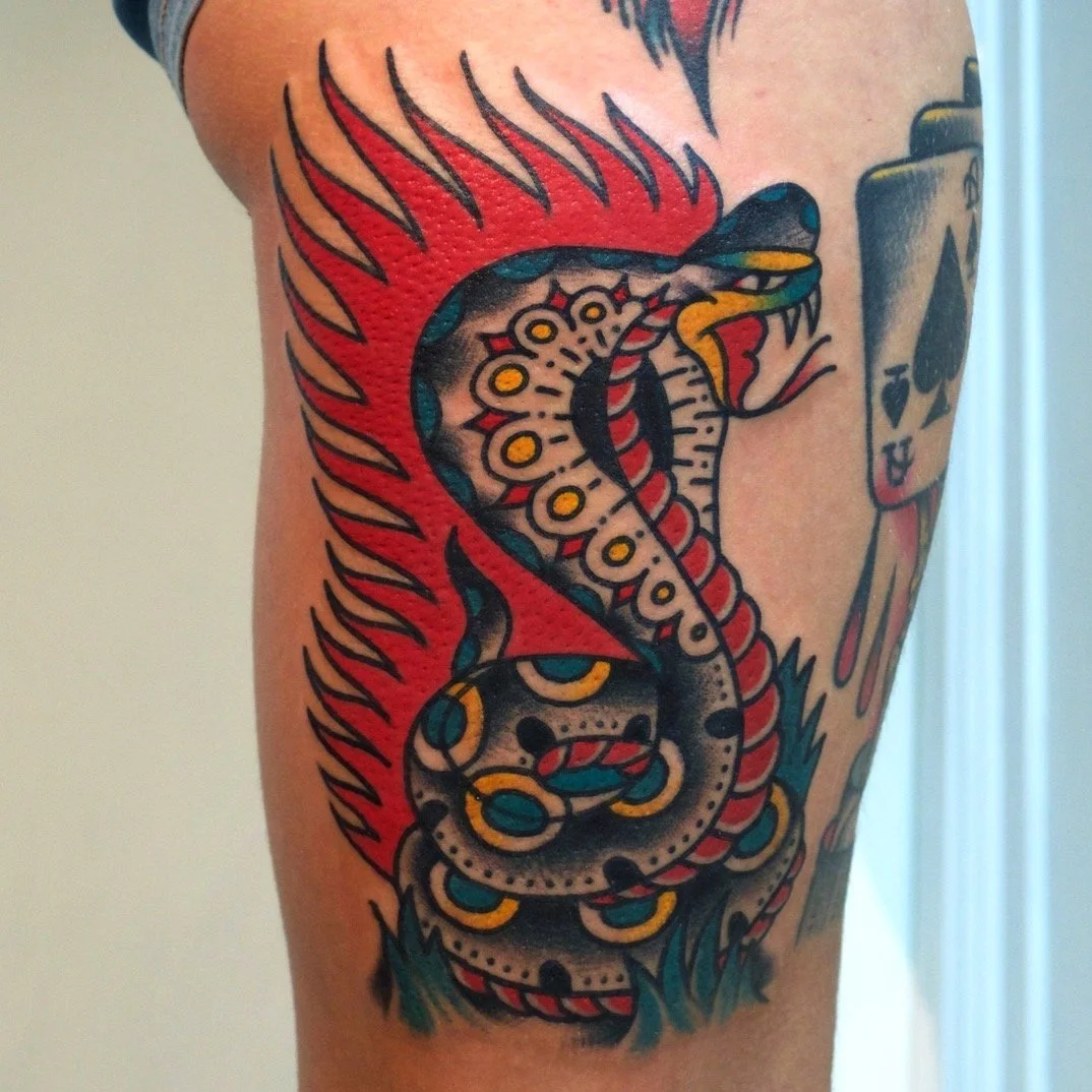 Traditional style tattoo of a coiled snake with red and black scales, open mouth, and tongue out, surrounded by flames and decorative elements.