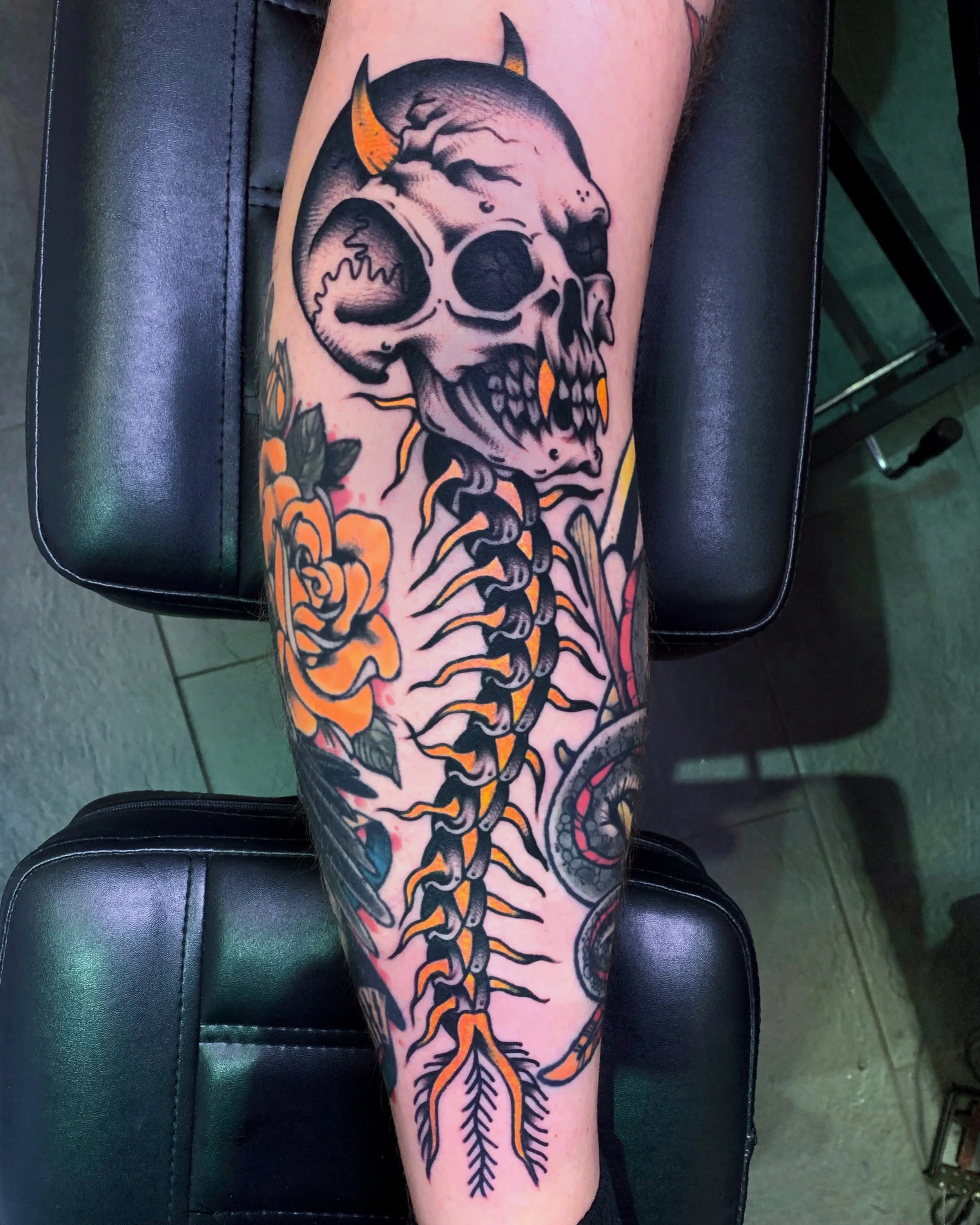 Tattoo of a skull with horns, floral elements, and a snake on someone's leg.