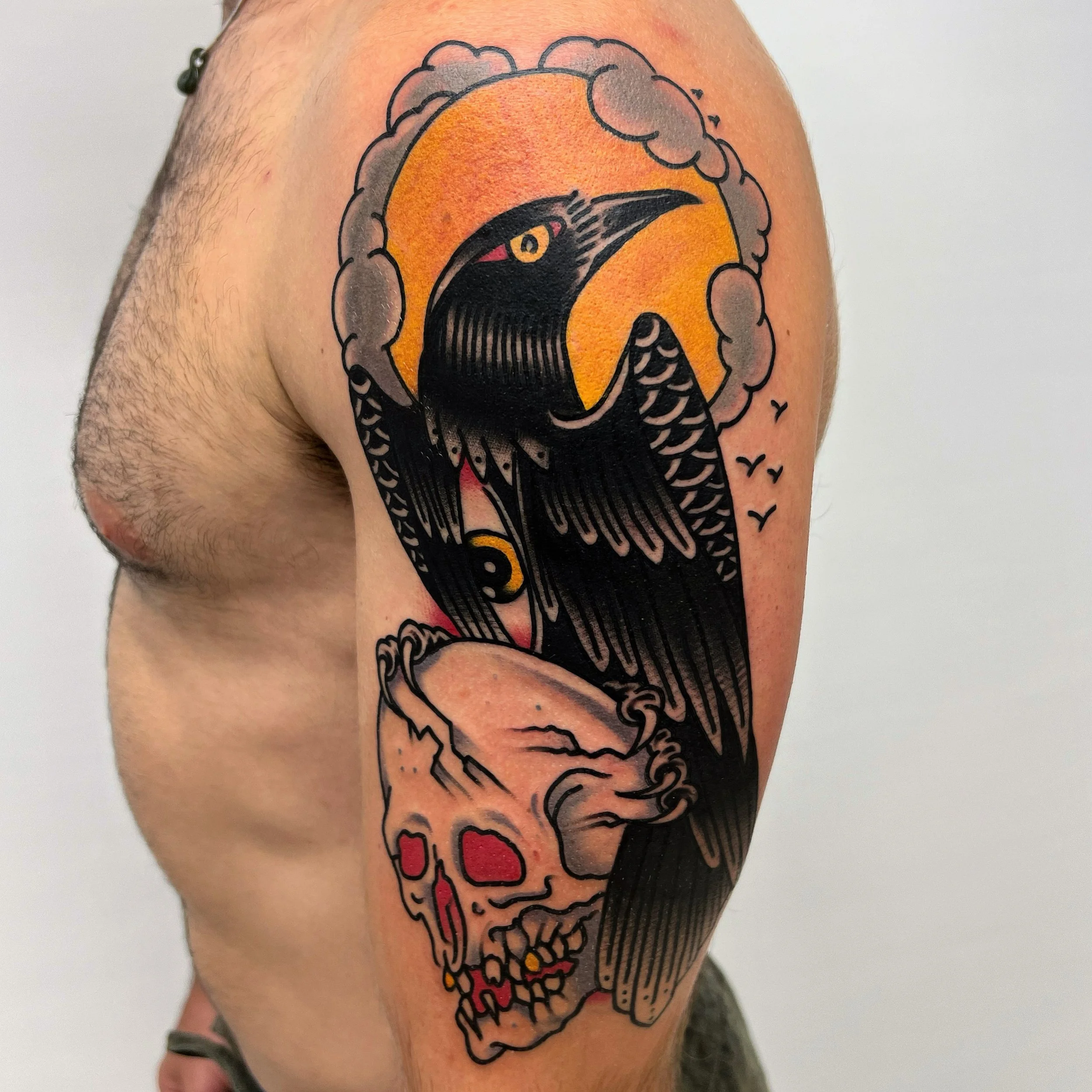Tattoo of a black crow with a yellow eye perched above a human skull, with a yellow sun or moon and clouds in the background, and small flying birds in the distance.