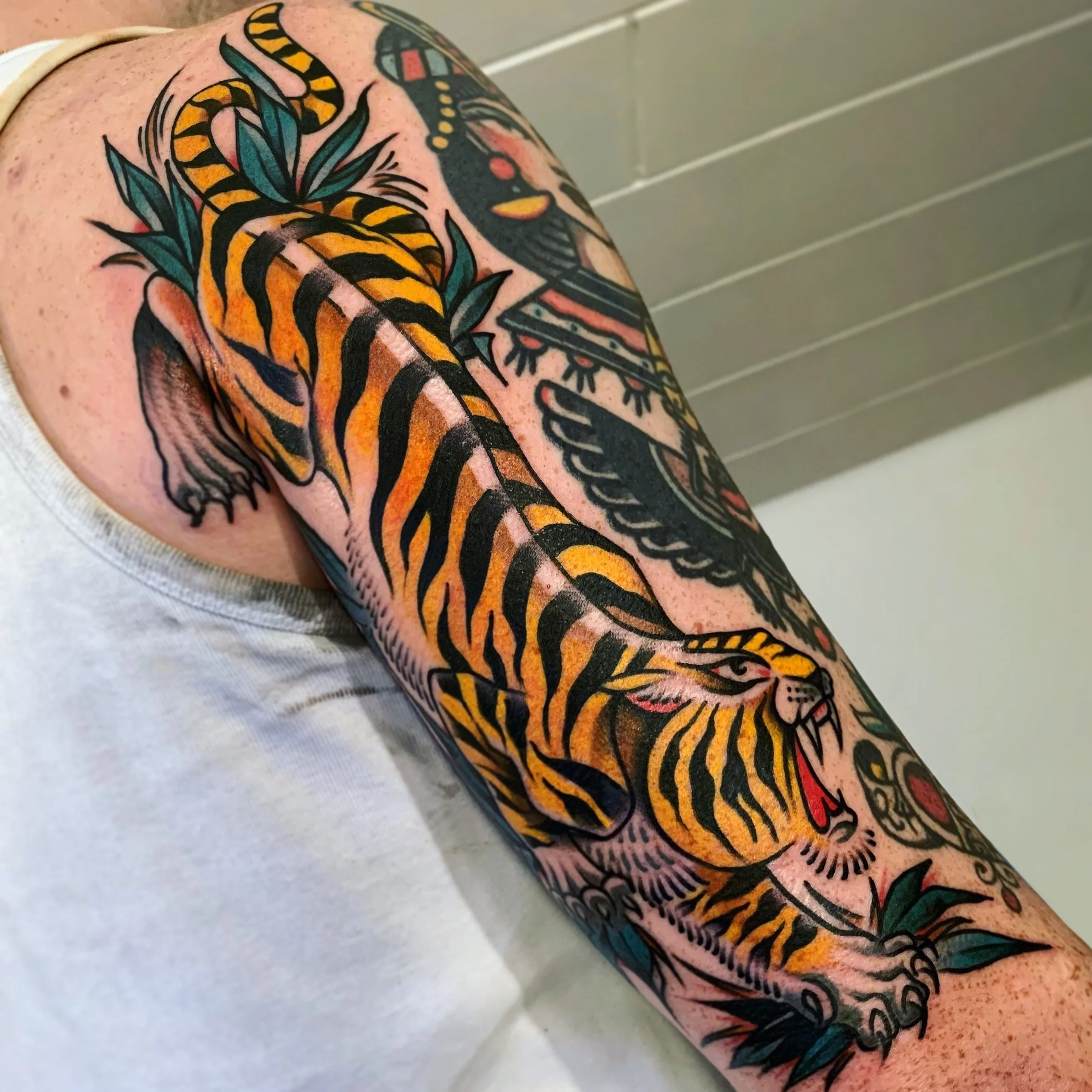 Colorful tattoo of a tiger with black stripes and orange fur, surrounded by green leaves and other decorative elements on a person's upper arm and shoulder.