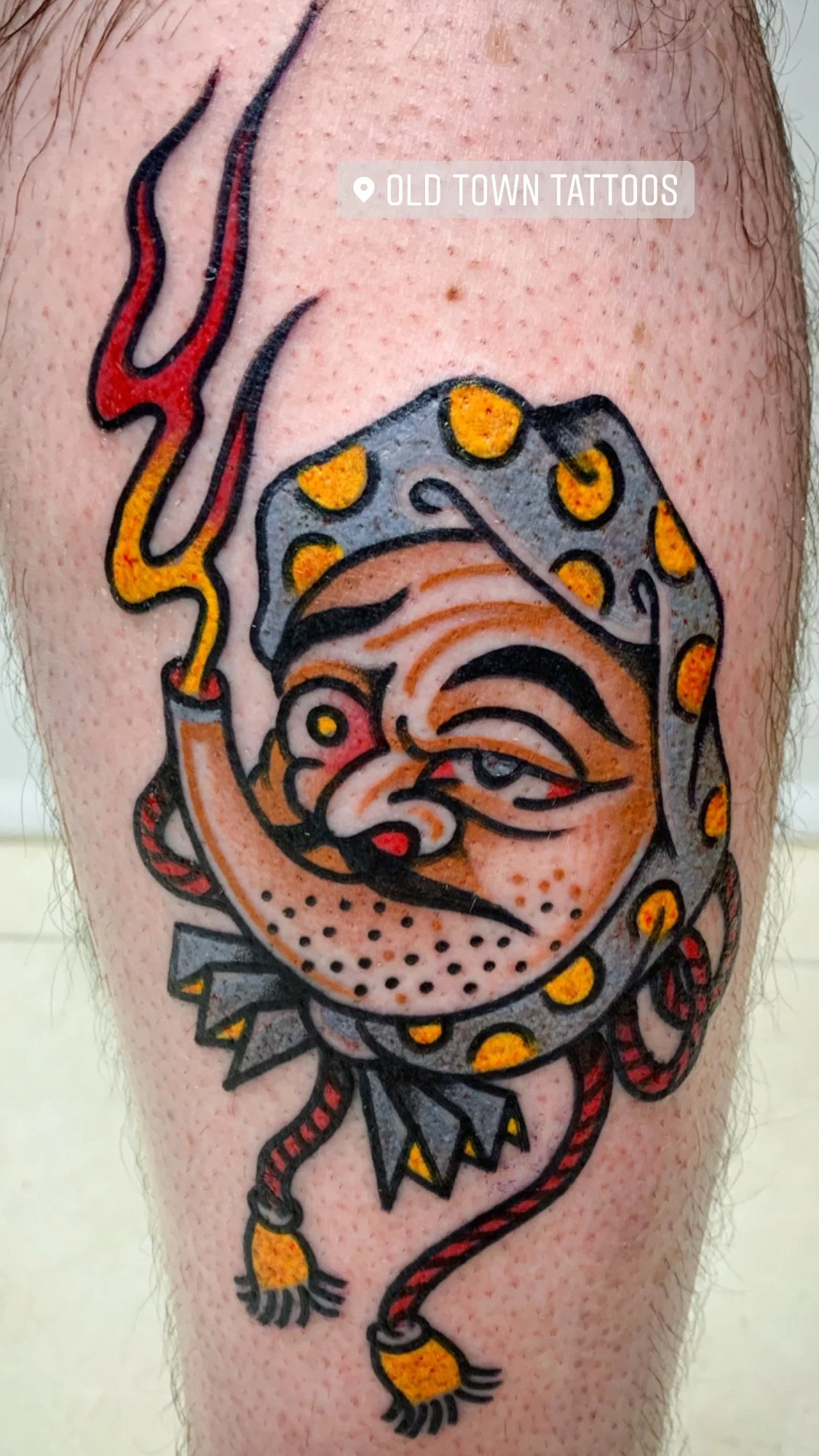 A colorful tattoo of a grumpy-looking old woman smoking a pipe, wearing a polka dot headscarf, located on the upper arm.