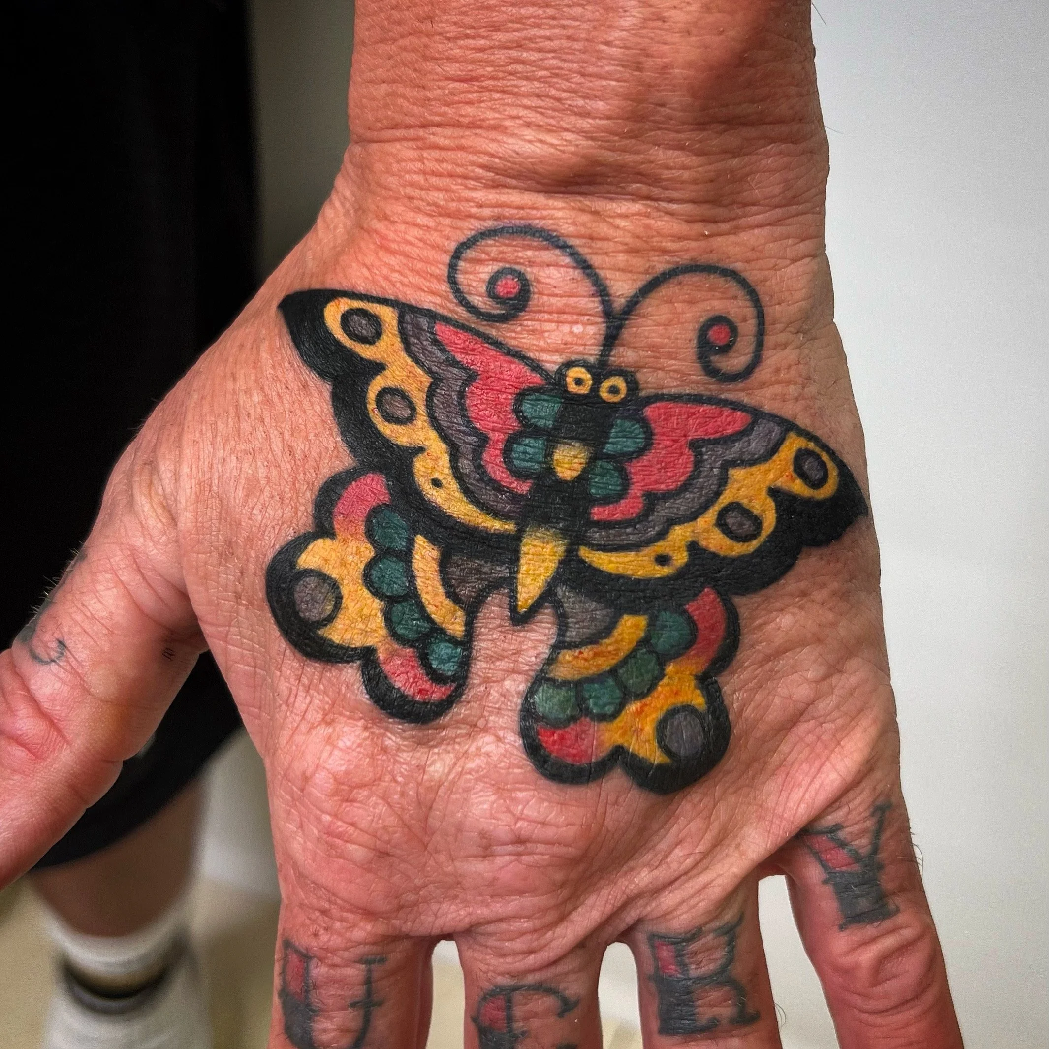 Colorful butterfly tattoo on a person's hand featuring yellow, red, green, black, and orange colors with intricate detailing and black outlines.
