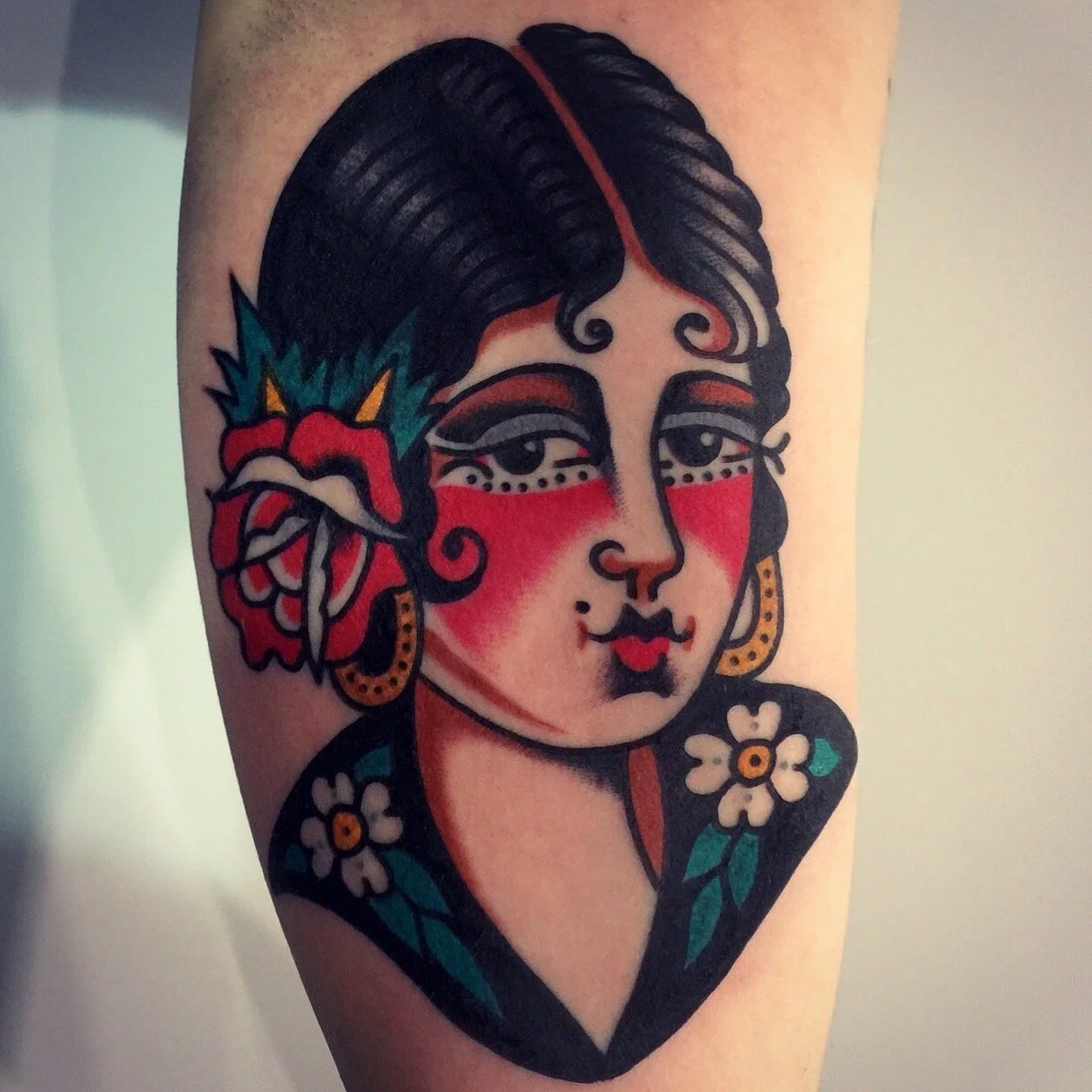 Traditional style tattoo of a woman with dark hair, red makeup, and floral accents, including a large red flower with green and yellow leaves behind her head