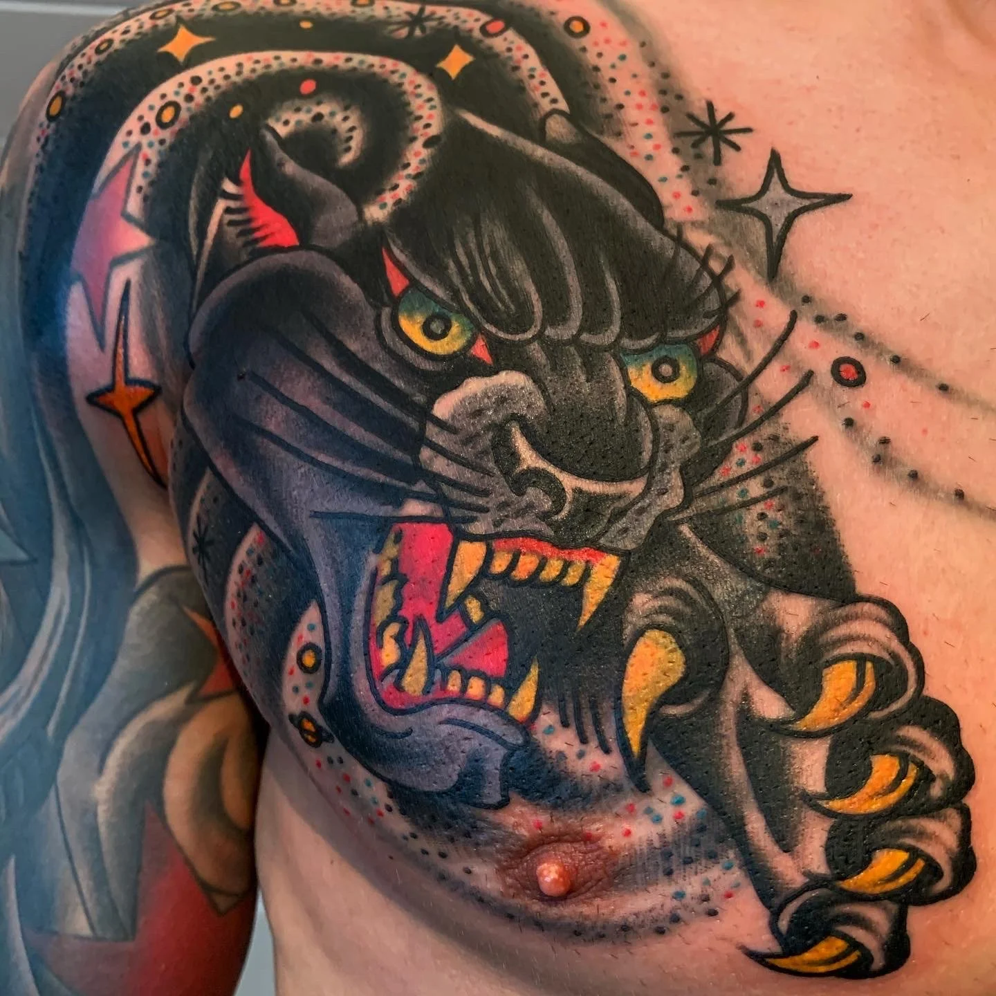 Tattoo of a fierce black panther with yellow eyes, sharp claws, and an open mouth showing teeth, on a person's chest.