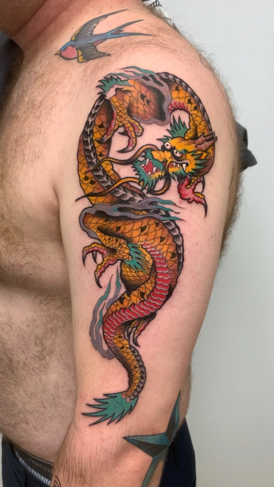 Colorful traditional Japanese-style dragon tattoo on man's upper arm with a small bird tattoo near shoulder.