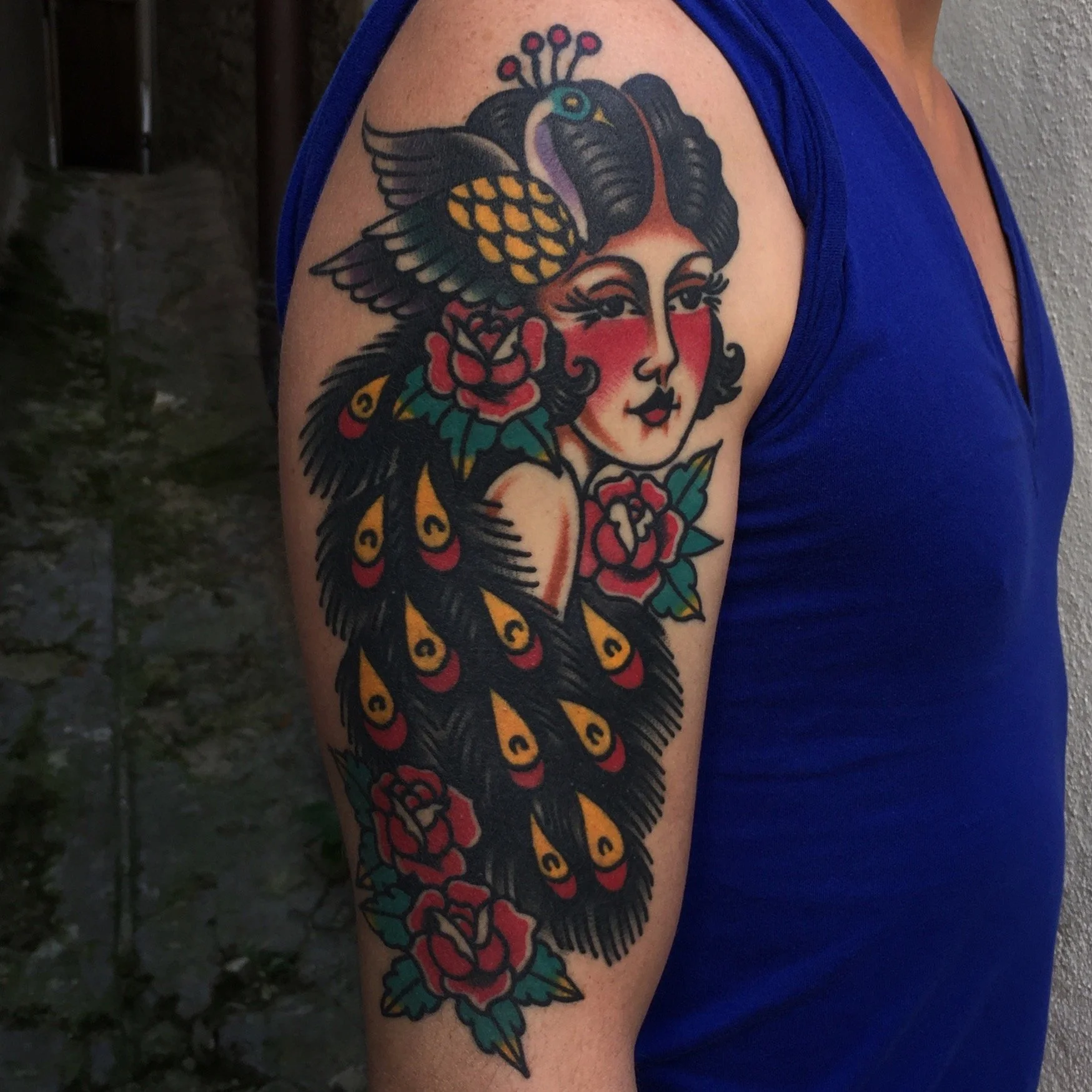 Tattoo of a stylized woman with roses, peacock feathers, and a bird on her head, on a person's upper arm.