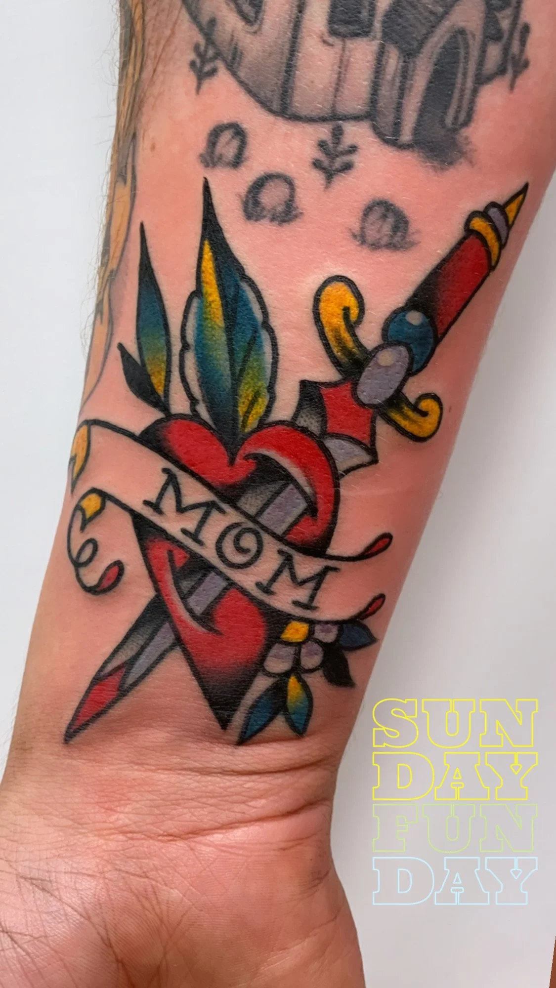 Tattoo of a dagger and colorful flowers with a banner that reads 'Mom' on person's arm. Text overlay says 'SUNDAY FUN DAY' in pastel colors.