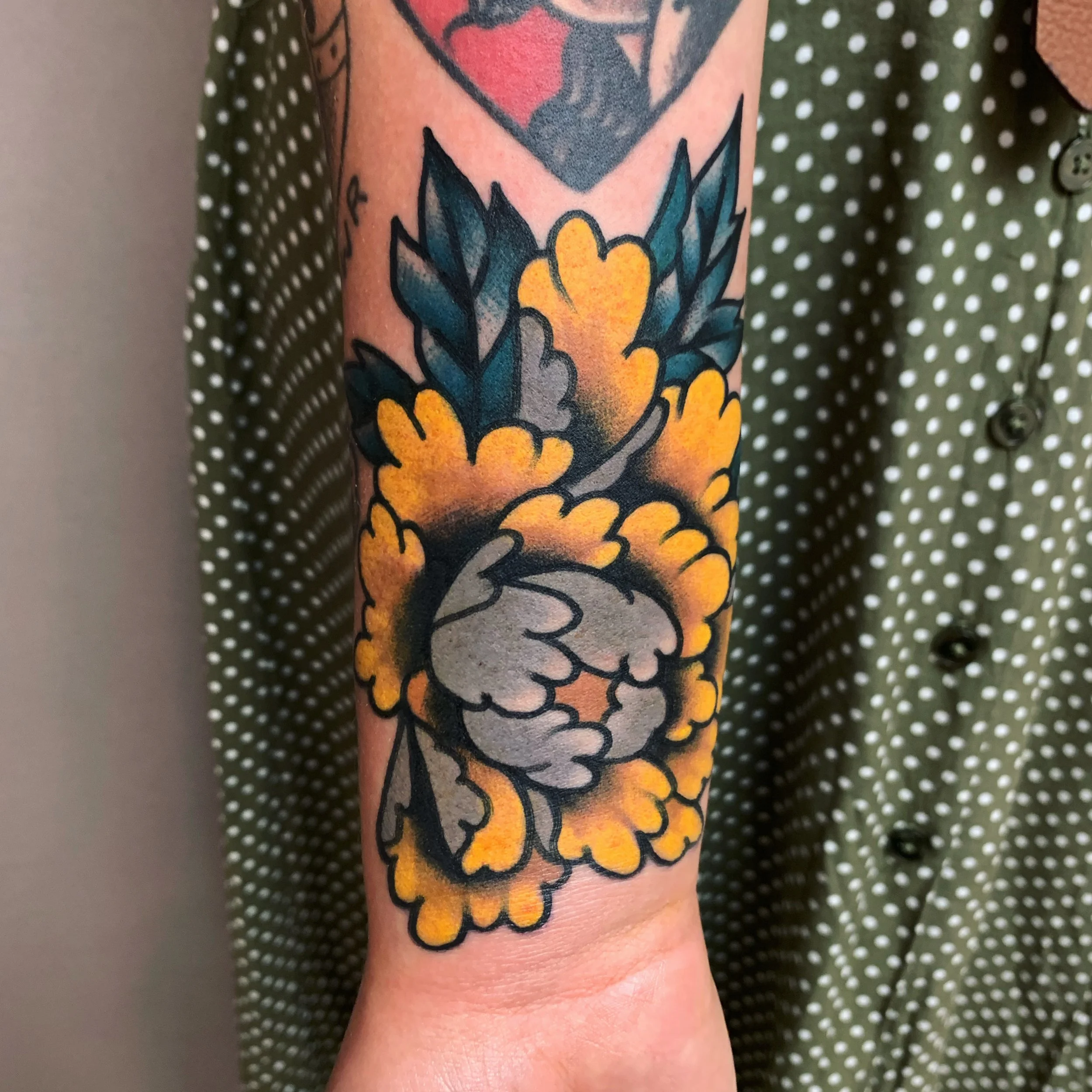Tattoo of a flower with yellow petals and dark green leaves on a person's forearm.