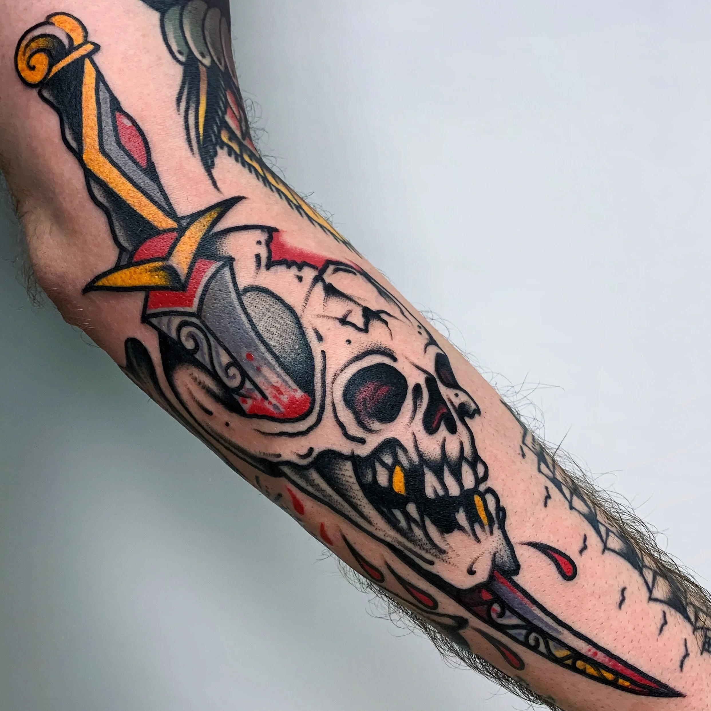 Tattoo of a skull with a sword through it on a person's arm, with black, red, yellow, and gray ink in a bold, traditional style.