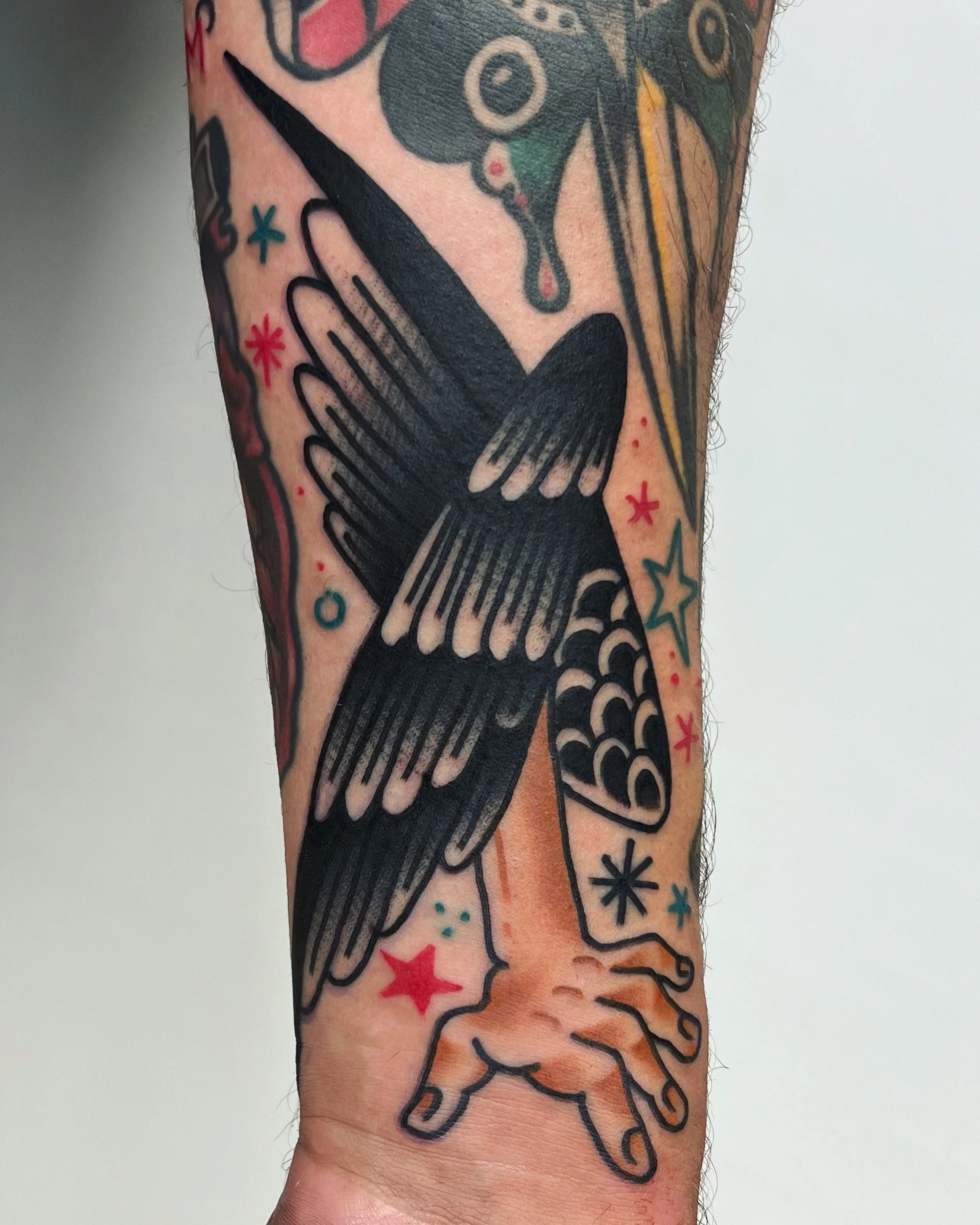 Tattoo of a black bird in flight with outstretched wings, perched on a person's hand, with colorful stars and abstract shapes surrounding it.