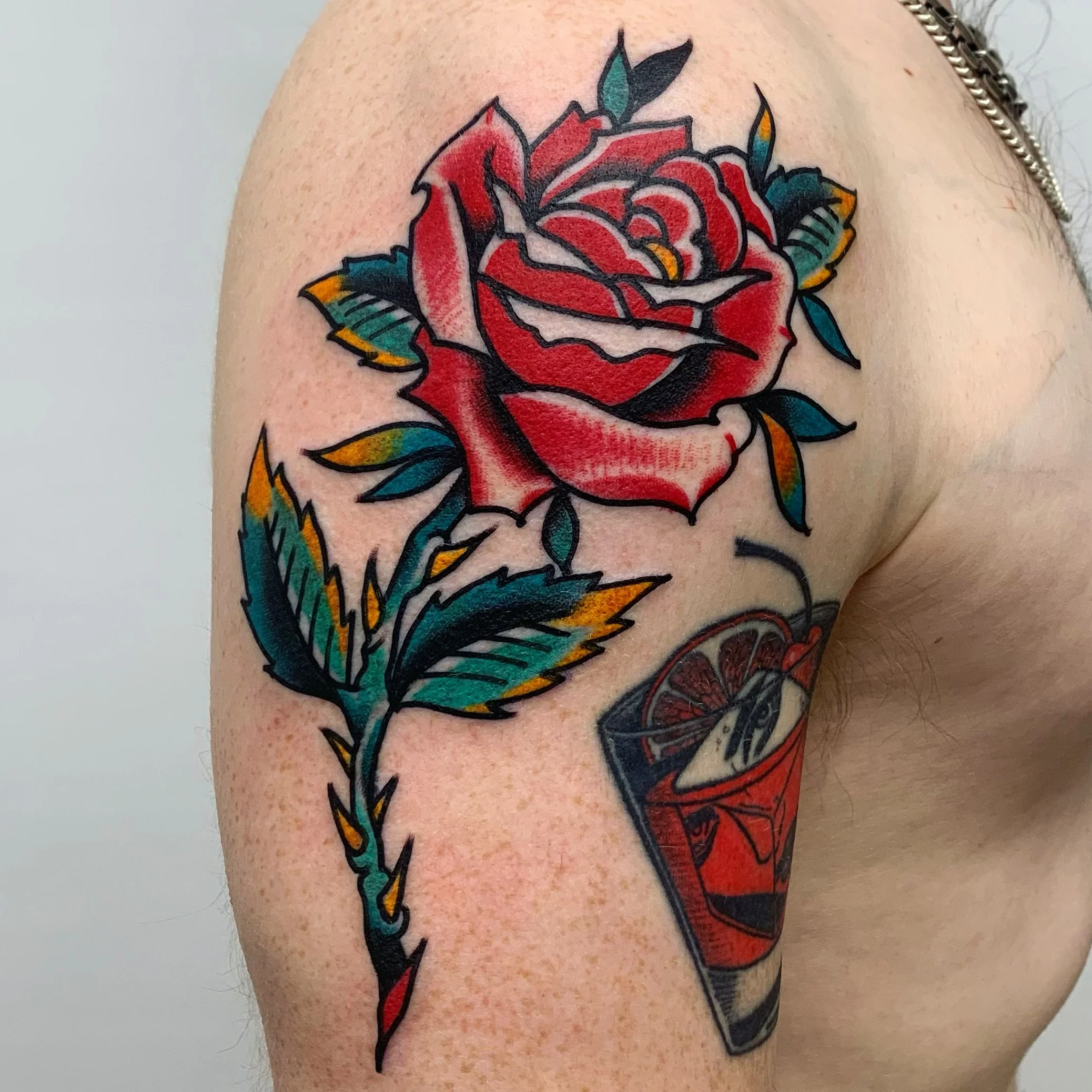 A colorful tattoo on a person's upper arm depicting a red rose with blue and yellow-tinted green leaves and a glass of a red drink with a lemon slice.