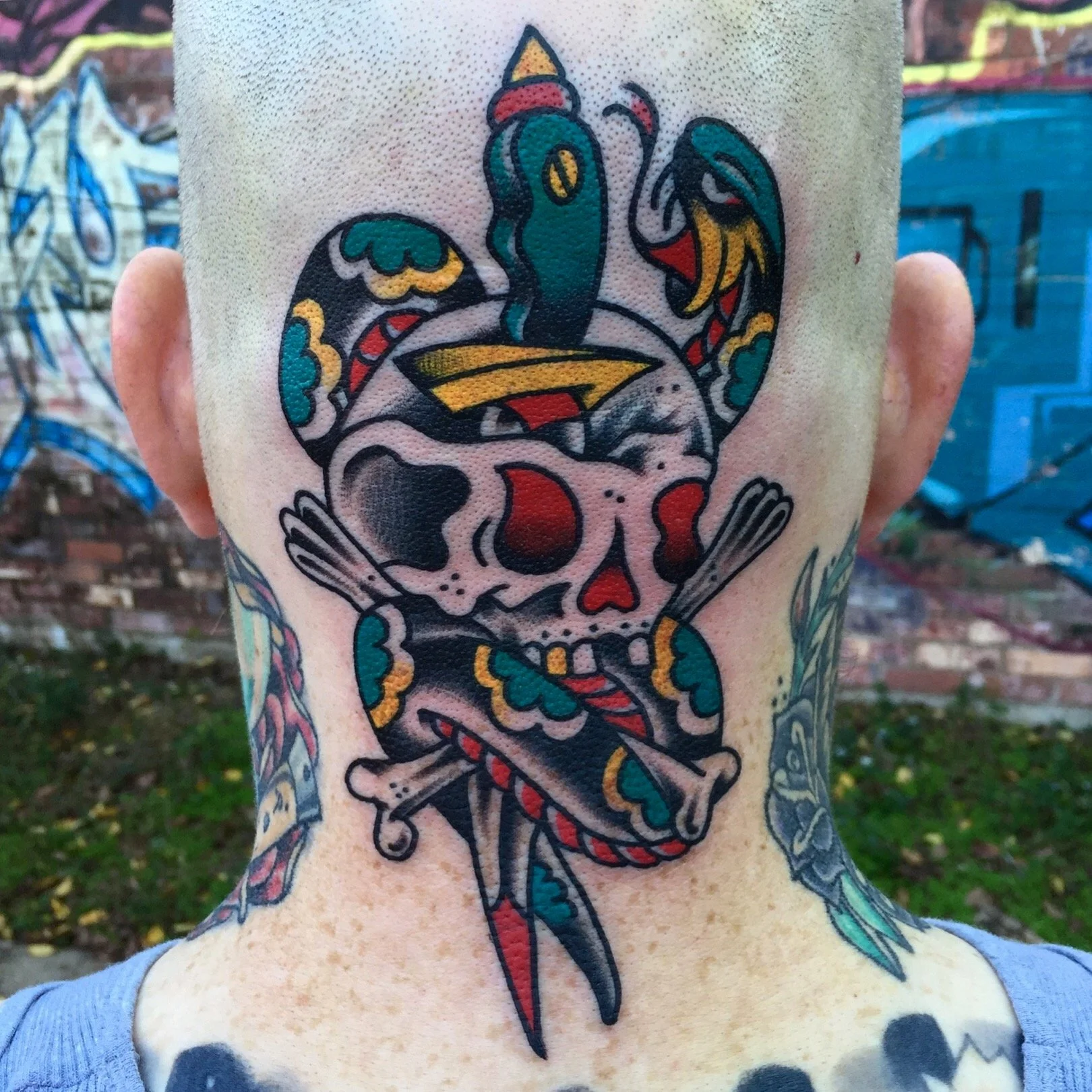 Tattoo of a colorful skull with a snake wrapping around it and a dagger through it, on the back of a person's neck.