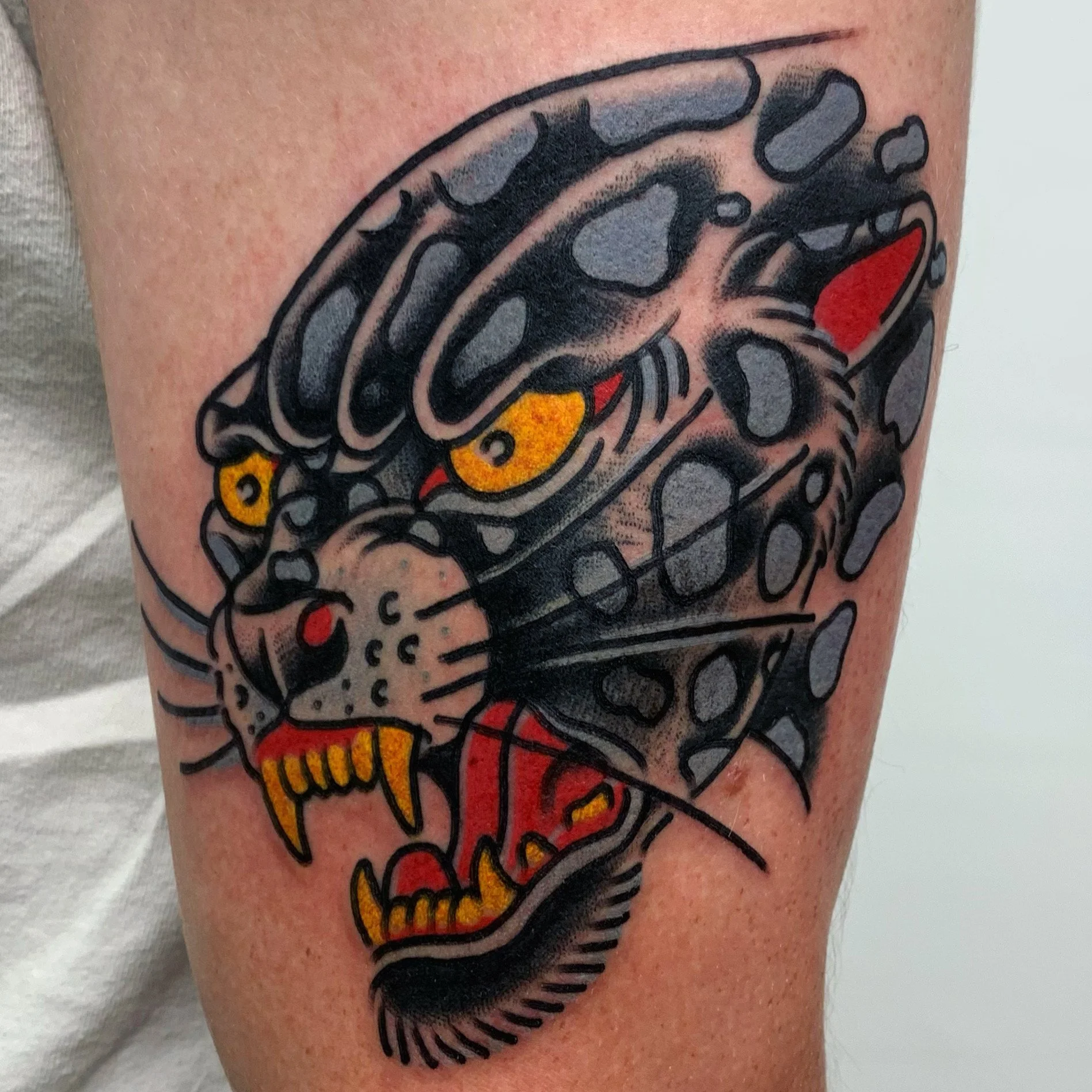 Tattoo of a fierce leopard head with yellow eyes, red tongue, and sharp teeth, designed in traditional tattoo style with bold black outlines and vibrant colors.