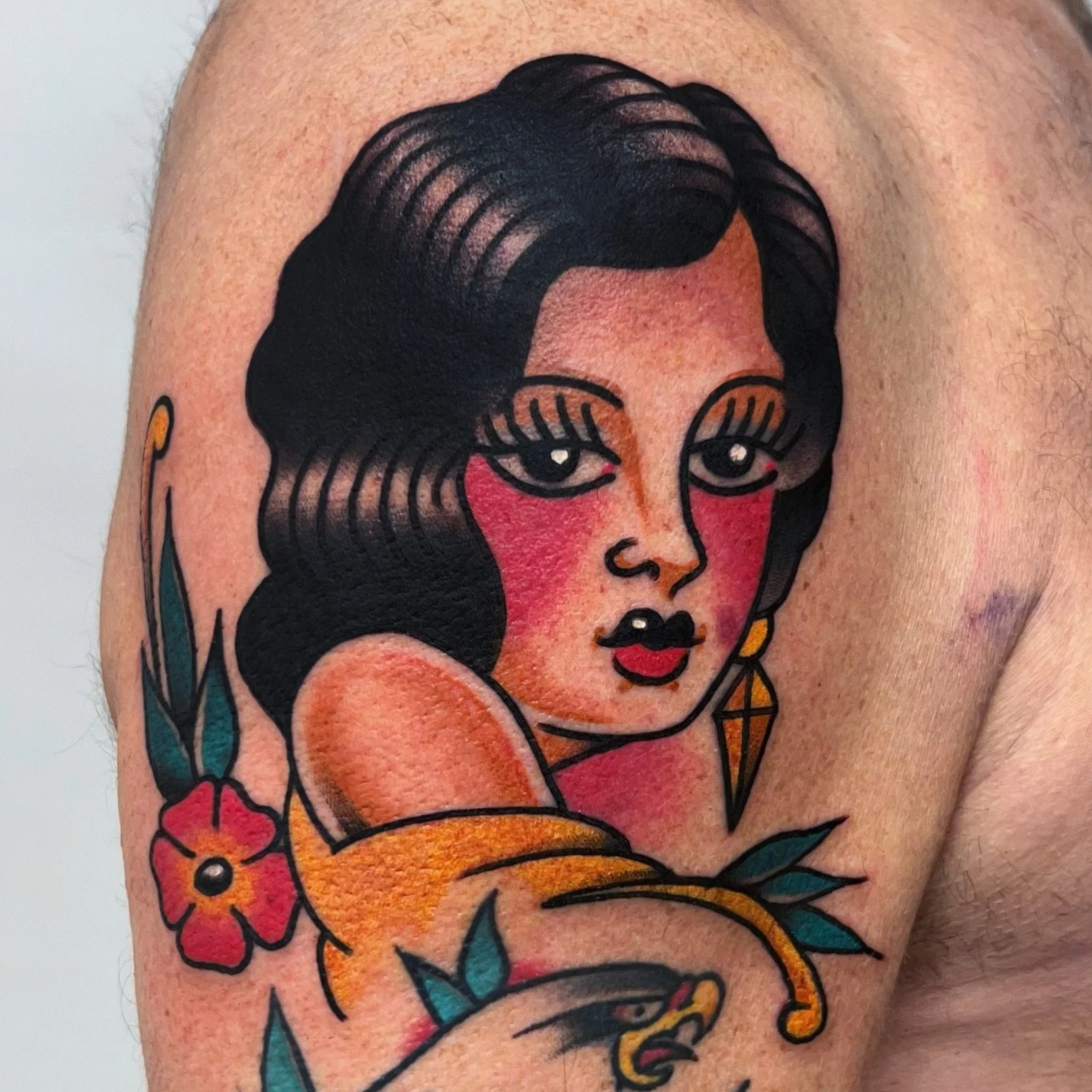 Tattoo of a stylized woman with dark hair, red cheeks, red lips, and gold earrings surrounded by a flower and a snake.