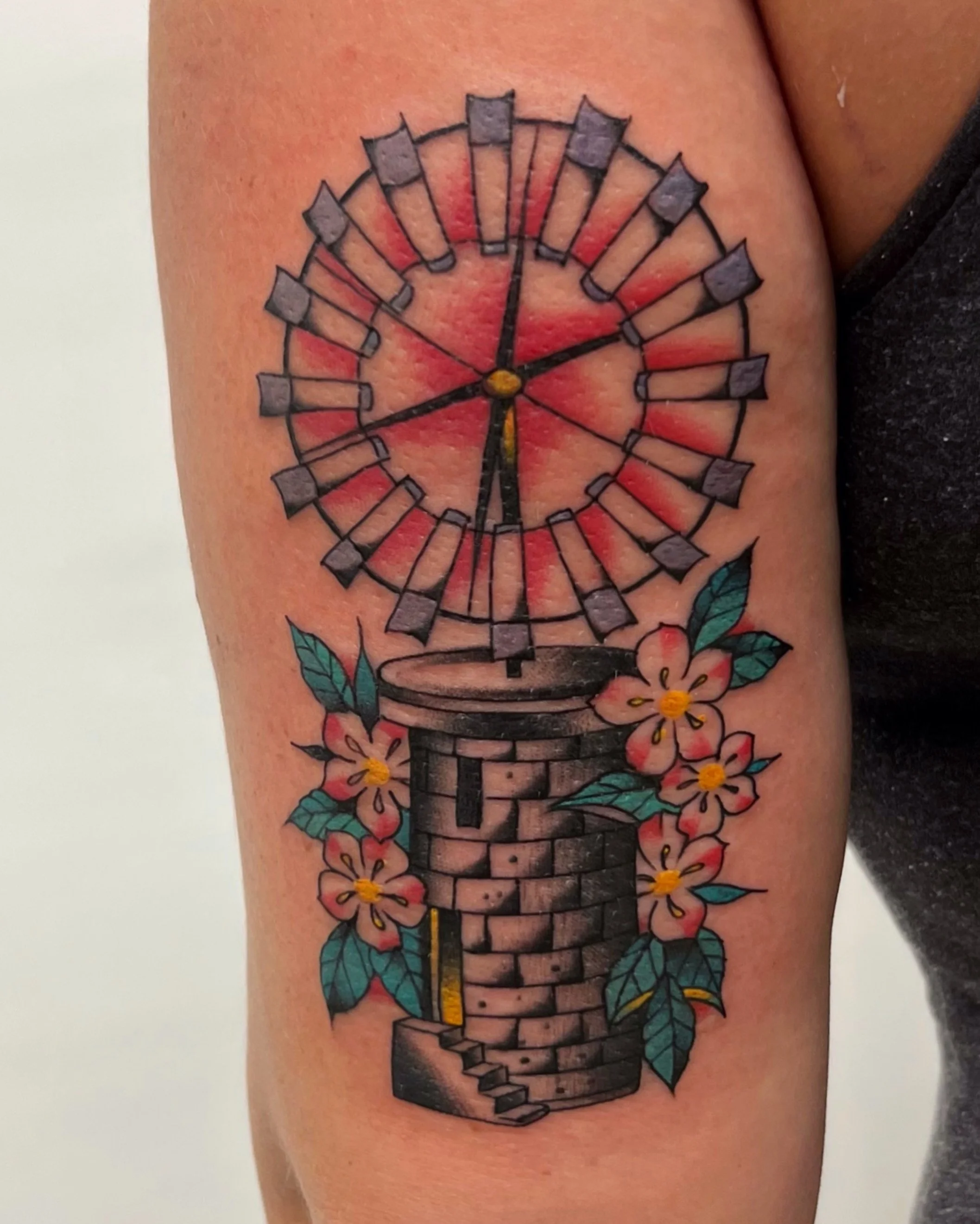Colorful tattoo of a Ferris wheel and a brick well with pink flowers and green leaves on a person's upper arm.
