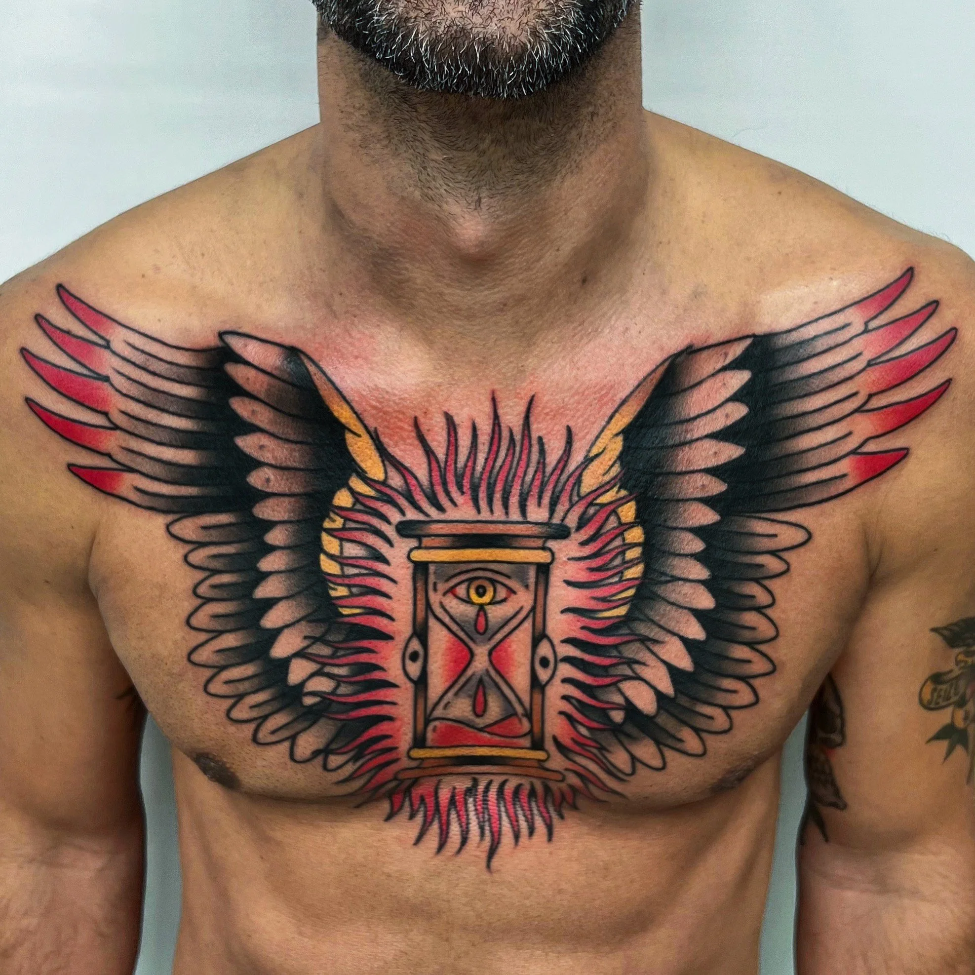 Tattoo of an eye within an hourglass, surrounded by wings on a man's chest.
