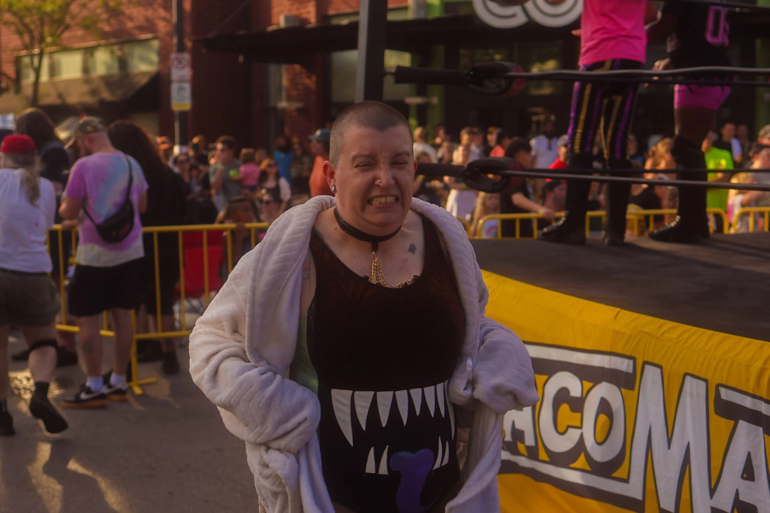 A person with a shaved head and tattoos on their neck, wearing a black dress with a large monster mouth graphic, and a white hoodie, squinting and making a grimace at a crowded outdoor event with many people in the background. WORK BY DAVEY