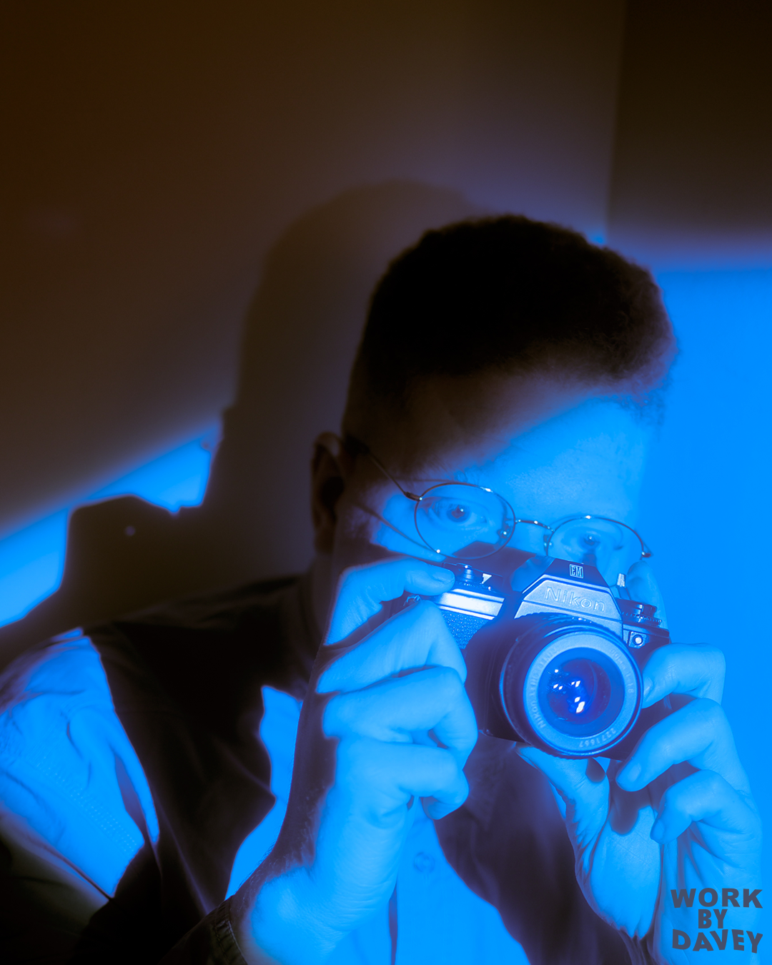 Person with glasses takes a photo with a Nikon camera, illuminated by blue light, with a shadow on the wall behind. WORK BY DAVEY