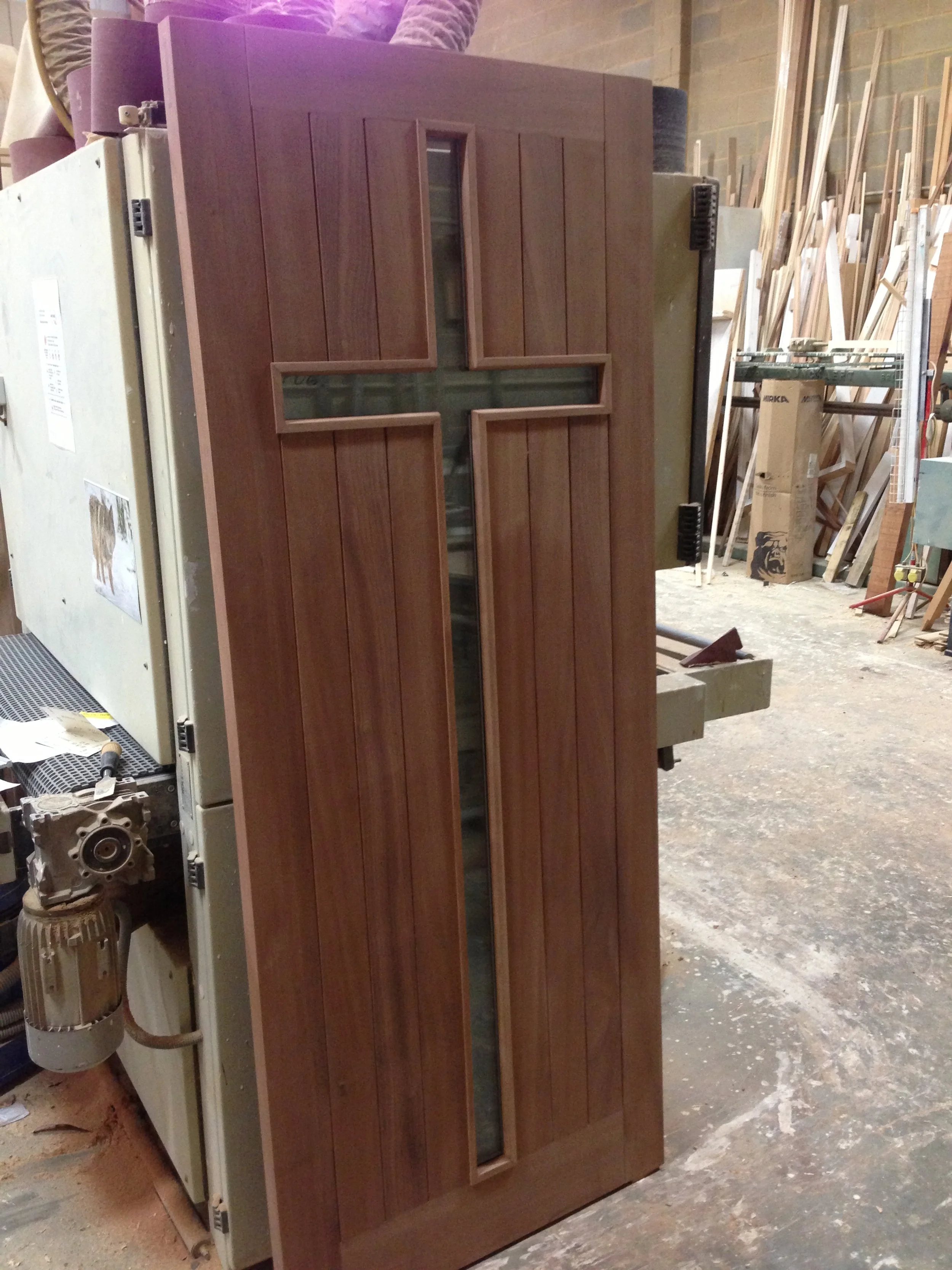 A wooden door with a cross-shaped glass panel in the center, situated in a woodworking shop.