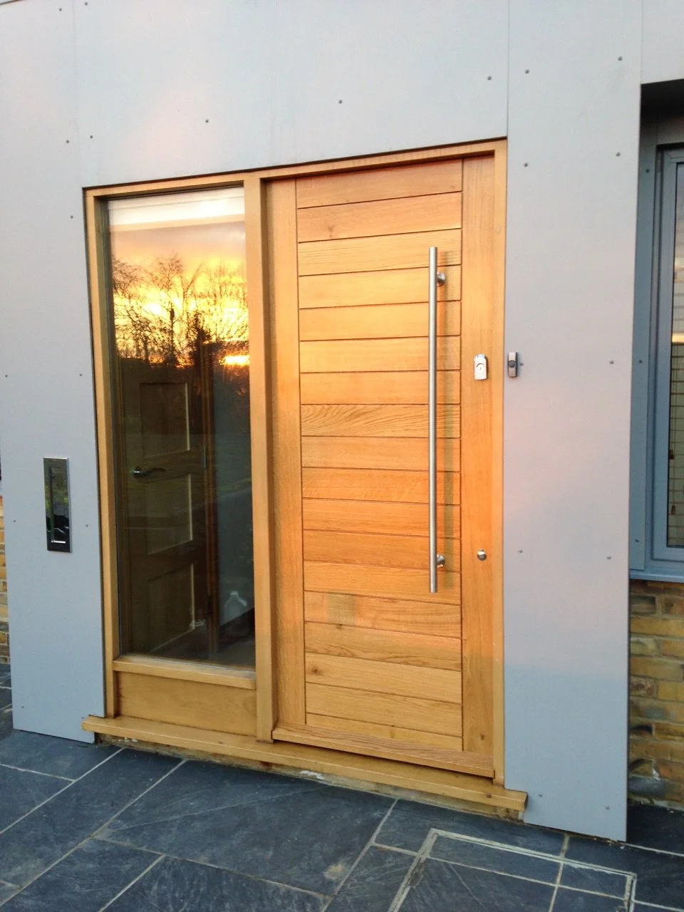 Modern exterior door with vertical wooden slats and a long metal handle, adjacent to a sidelight window reflecting a sunset and trees.