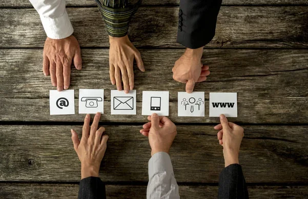 Hands reaching for icons representing at, phone, email, mobile, people, and web on a wooden table.