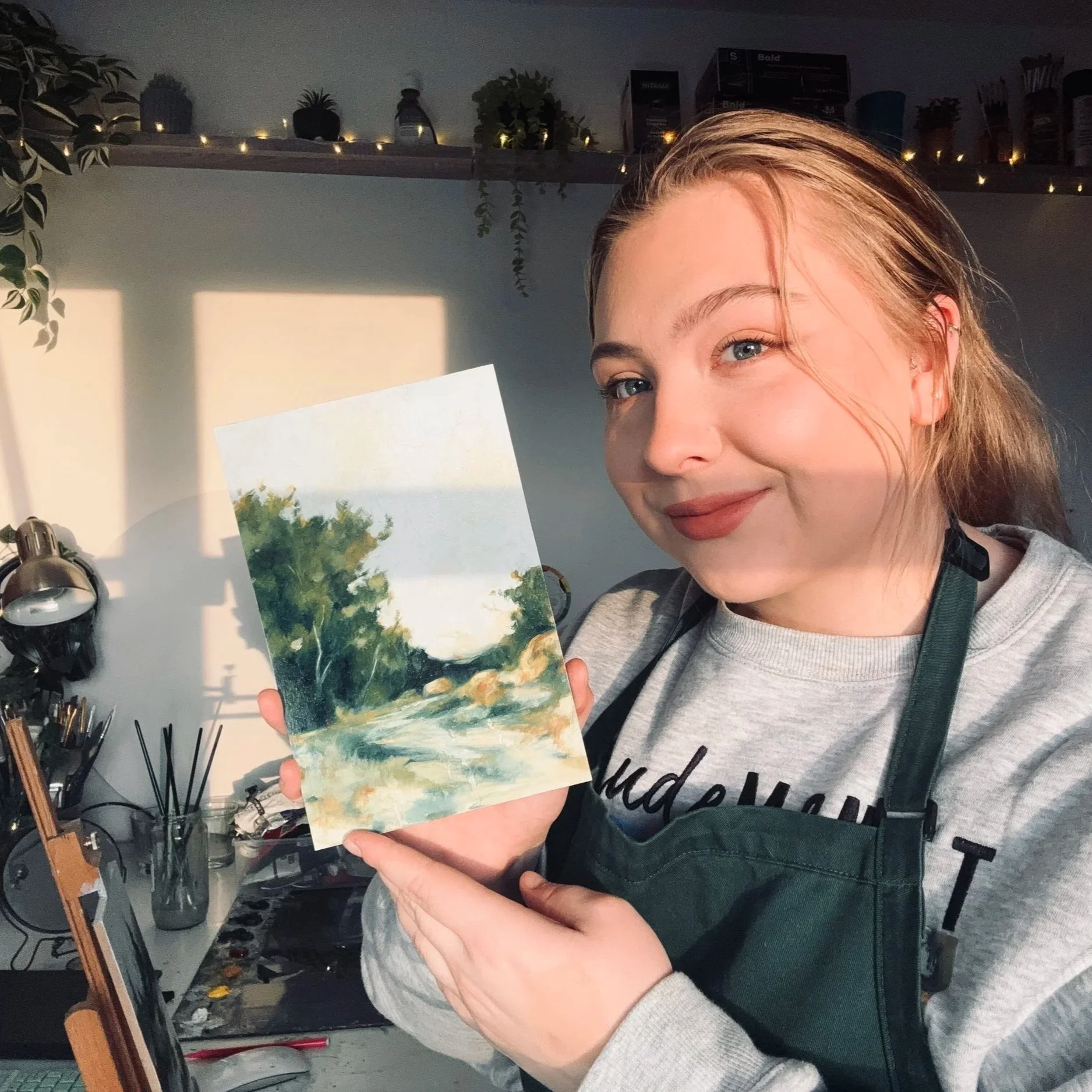 A woman with light skin and long blonde hair, wearing a gray shirt and a dark green apron, is smiling and holding up a small watercolor landscape painting of a river with trees. She is in an art studio with sunlight casting shadows on the wall behind her, and art supplies like brushes, paints, and a palette are visible on her workspace.