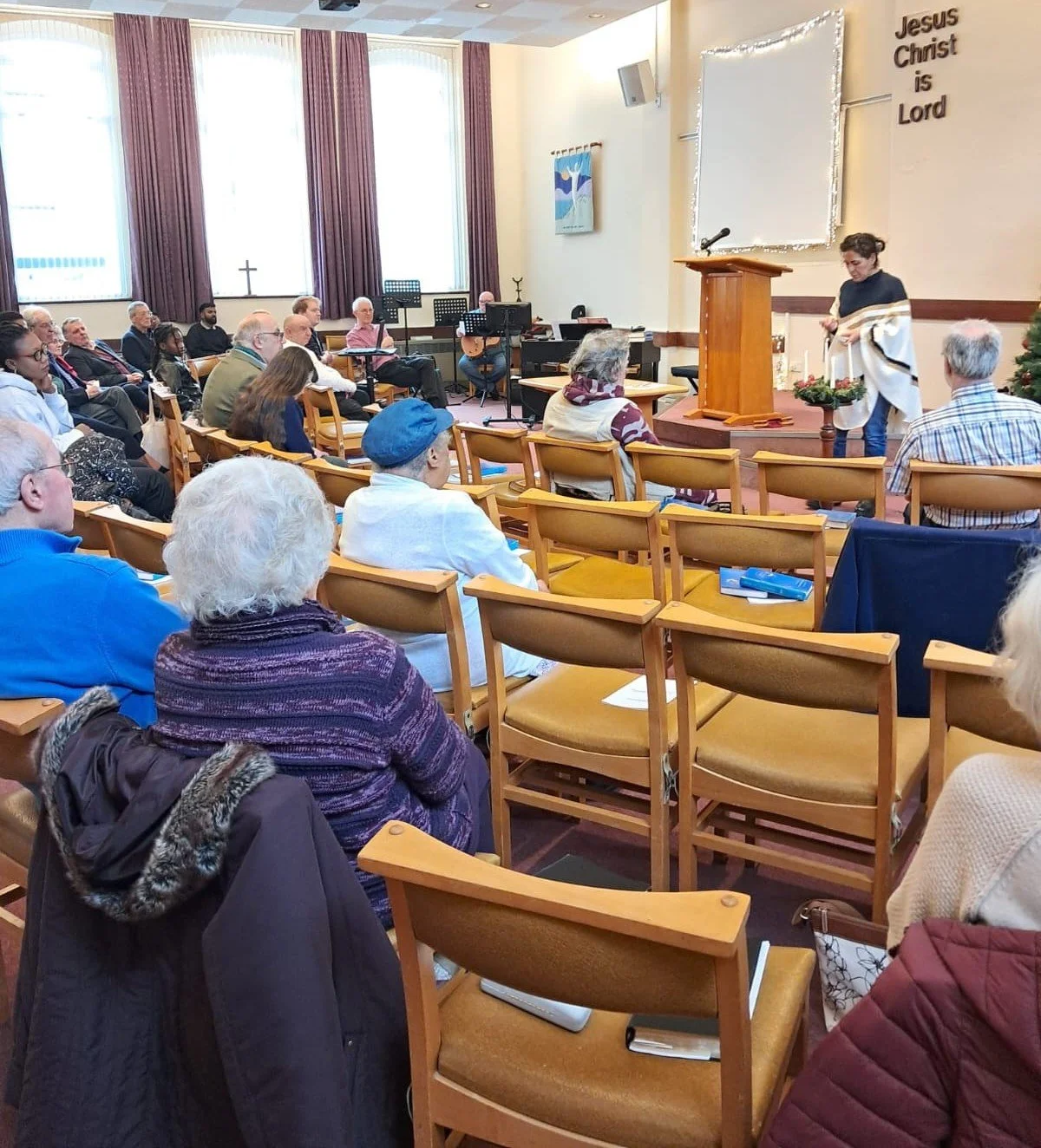 A diverse group of people attending a church service inside a church. There is a preacher at the podium and a sign that says 'Jesus Christ is Lord' on the wall.