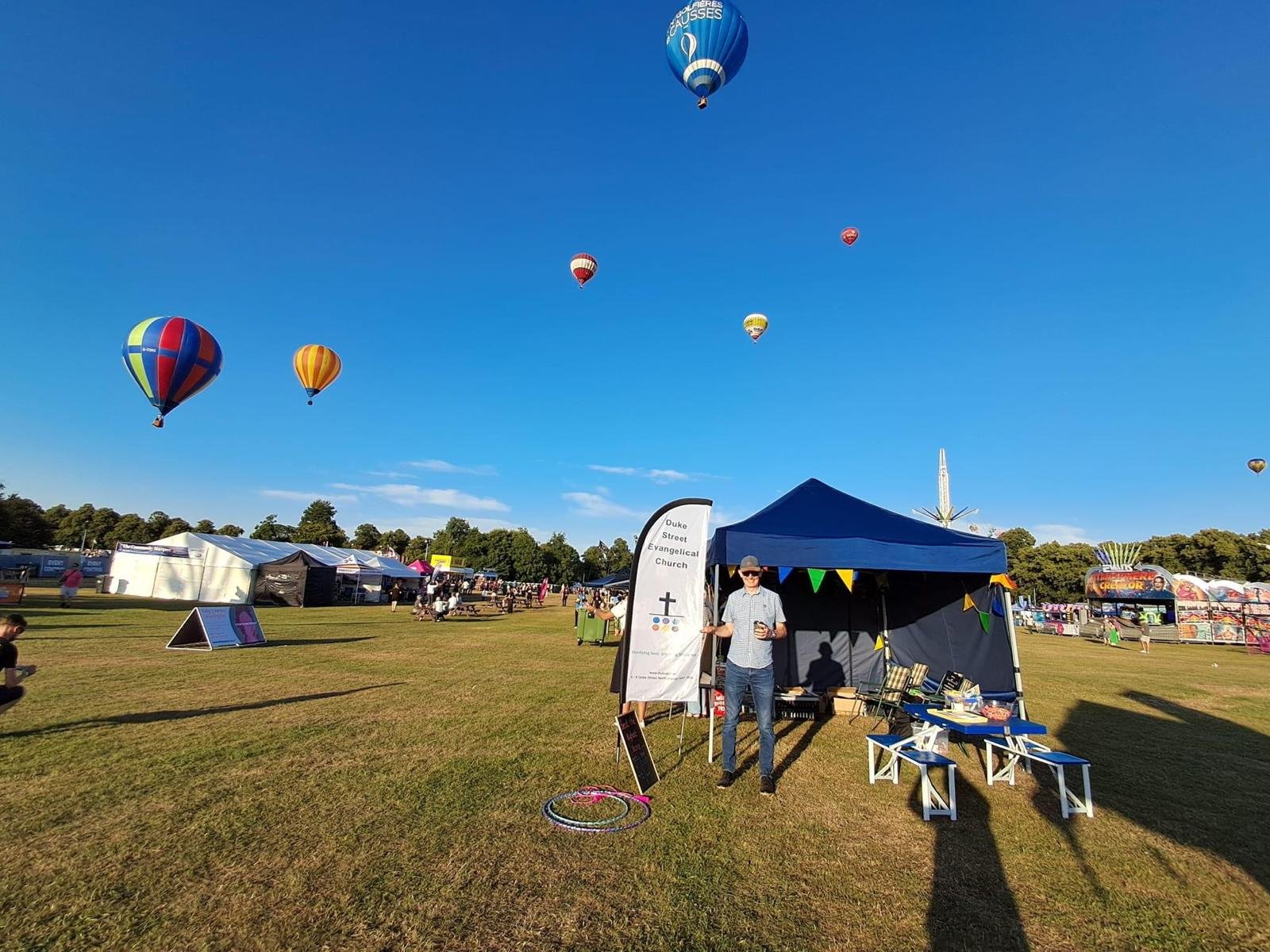 The church stand at the Northampton Balloon festival, with hot air balloons flying in a clear blue sky, and amusement rides in the background.