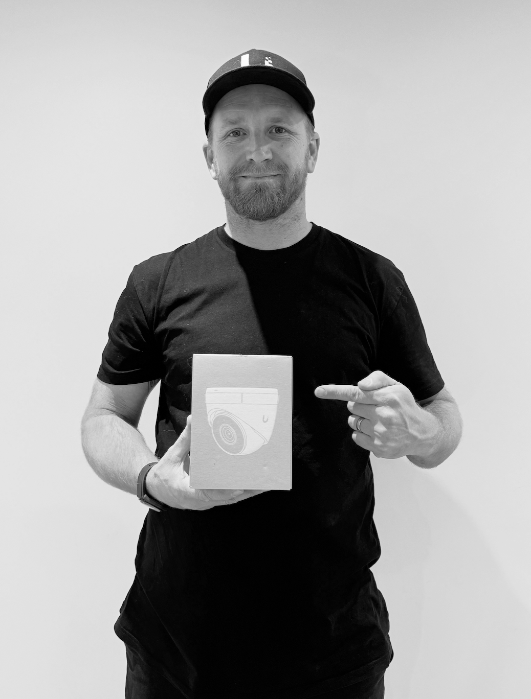 A man with a beard wearing a black t-shirt and a baseball cap, smiling and pointing at a box he is holding. The box has a spiral design on it.