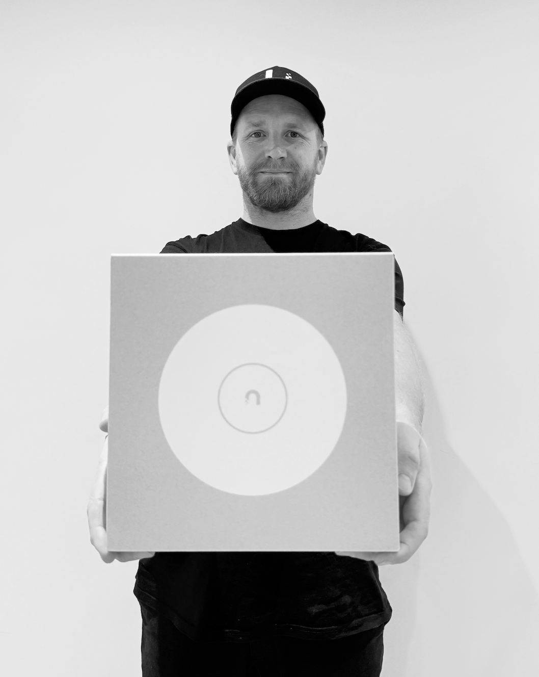 Man with a beard wearing a cap and black t-shirt holding a circular speaker or device with a minimalist design.