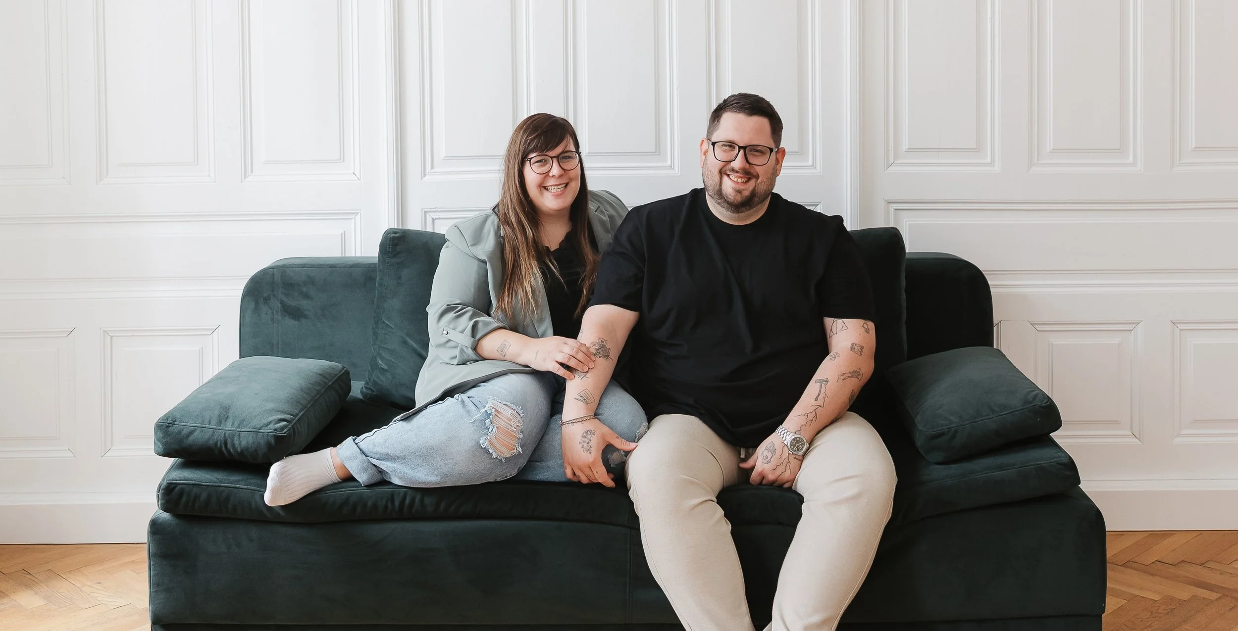 Ein lächelndes Paar sitzt auf einer dunklen Couch vor einer weißen Wand mit dekorativen Paneelen, der Mann trägt eine schwarze T-Shirt und beige Hosen, die Frau trägt eine graue Jacke und Jeans mit Rissen.