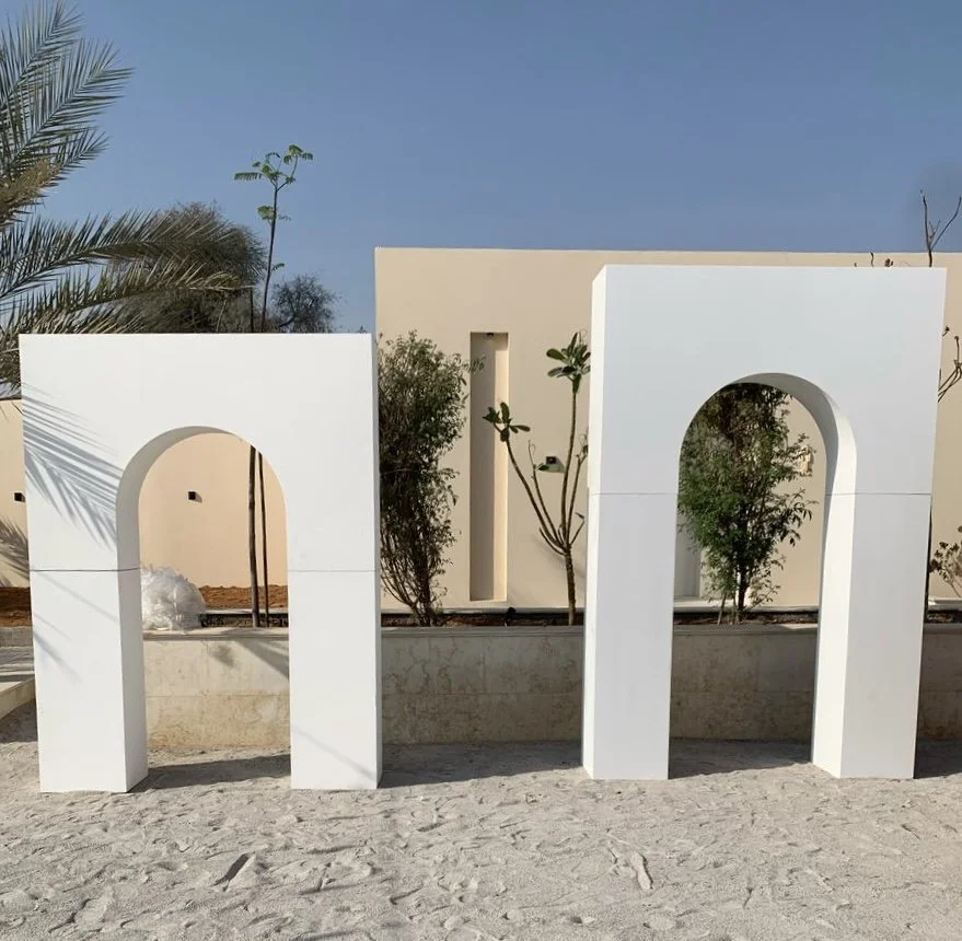 Large white sculptural letters spelling 'M' placed outdoors on sandy ground with trees and a beige wall in the background.