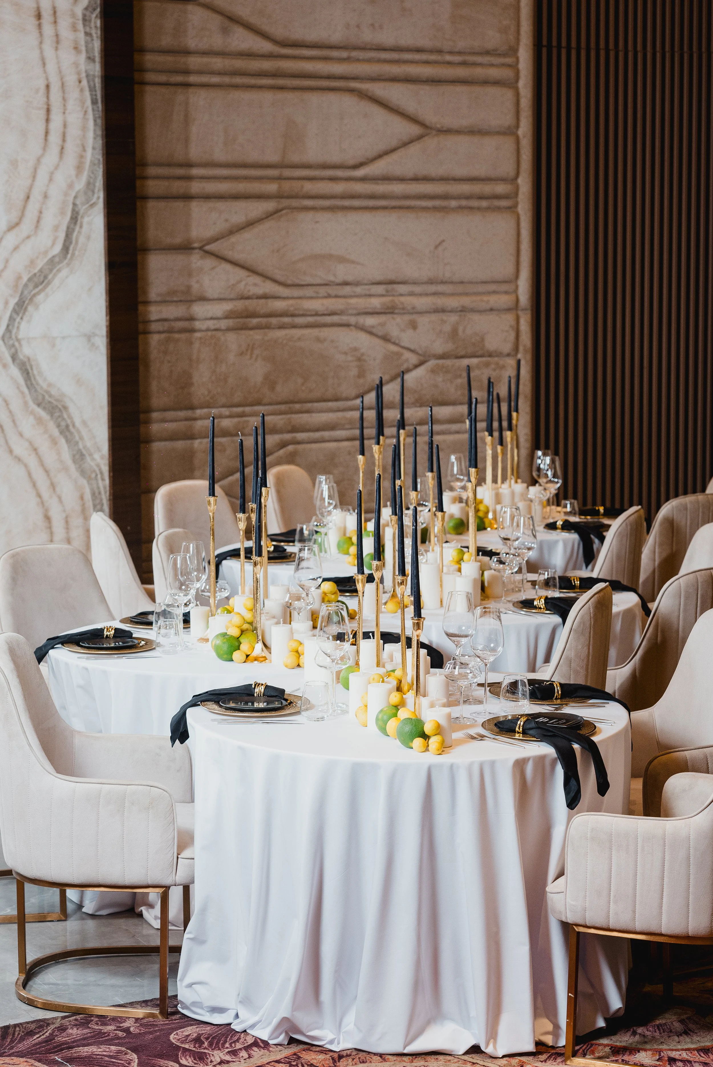 Elegant dining table set with white tablecloths, black napkins, gold cutlery, wine glasses, decorative candles, and fruit centerpieces featuring limes and yellow berries, in a luxurious room with beige upholstered chairs and textured walls.