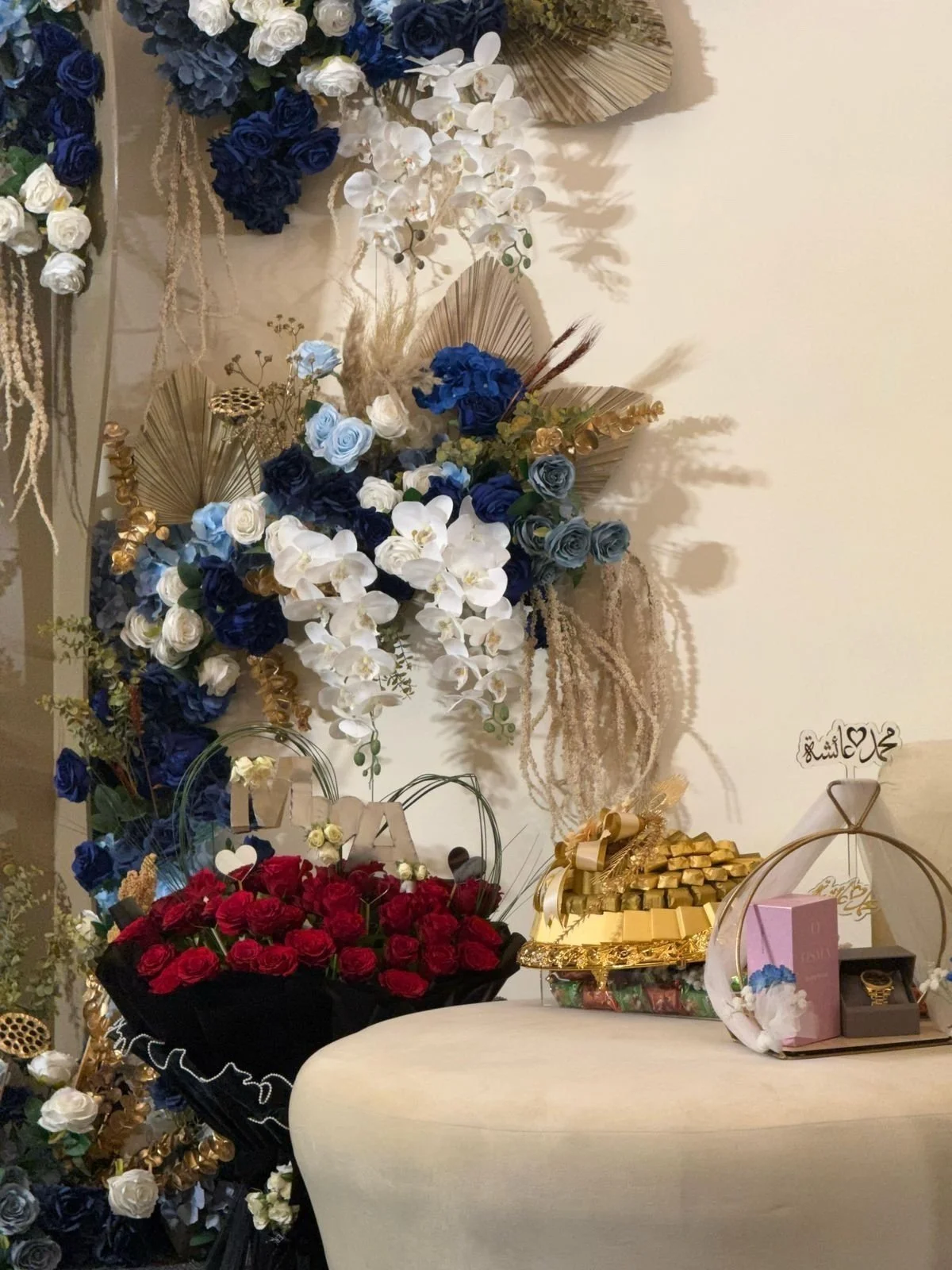 Decorative floral arrangement with white, blue, and red roses, gold accents, and gift boxes on a table.