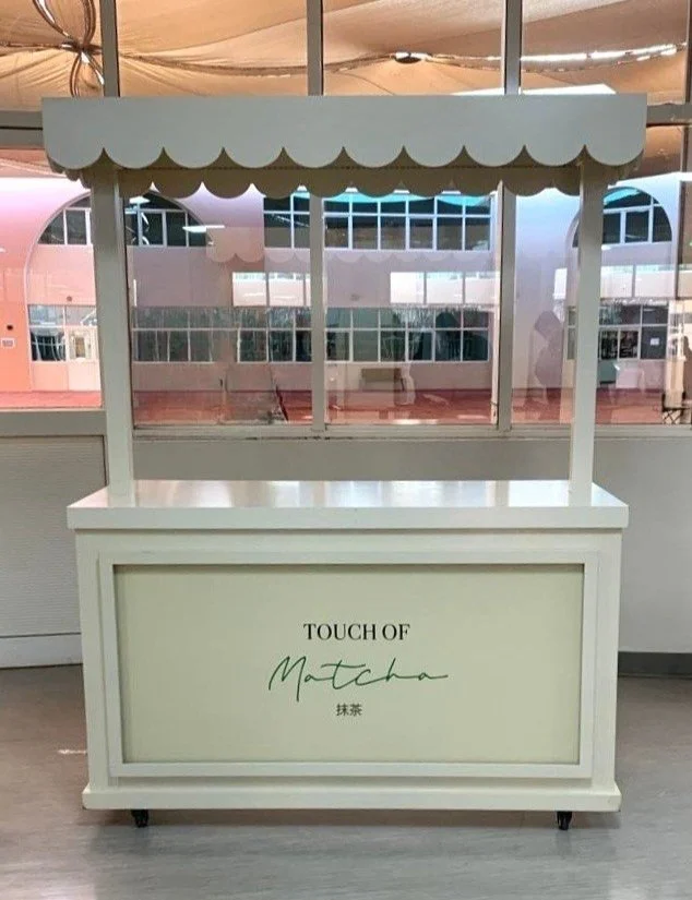 A white beverage stand with a scalloped canopy on top, labeled 'Touch of Matcha' in black and green text, situated indoors with large windows in the background.