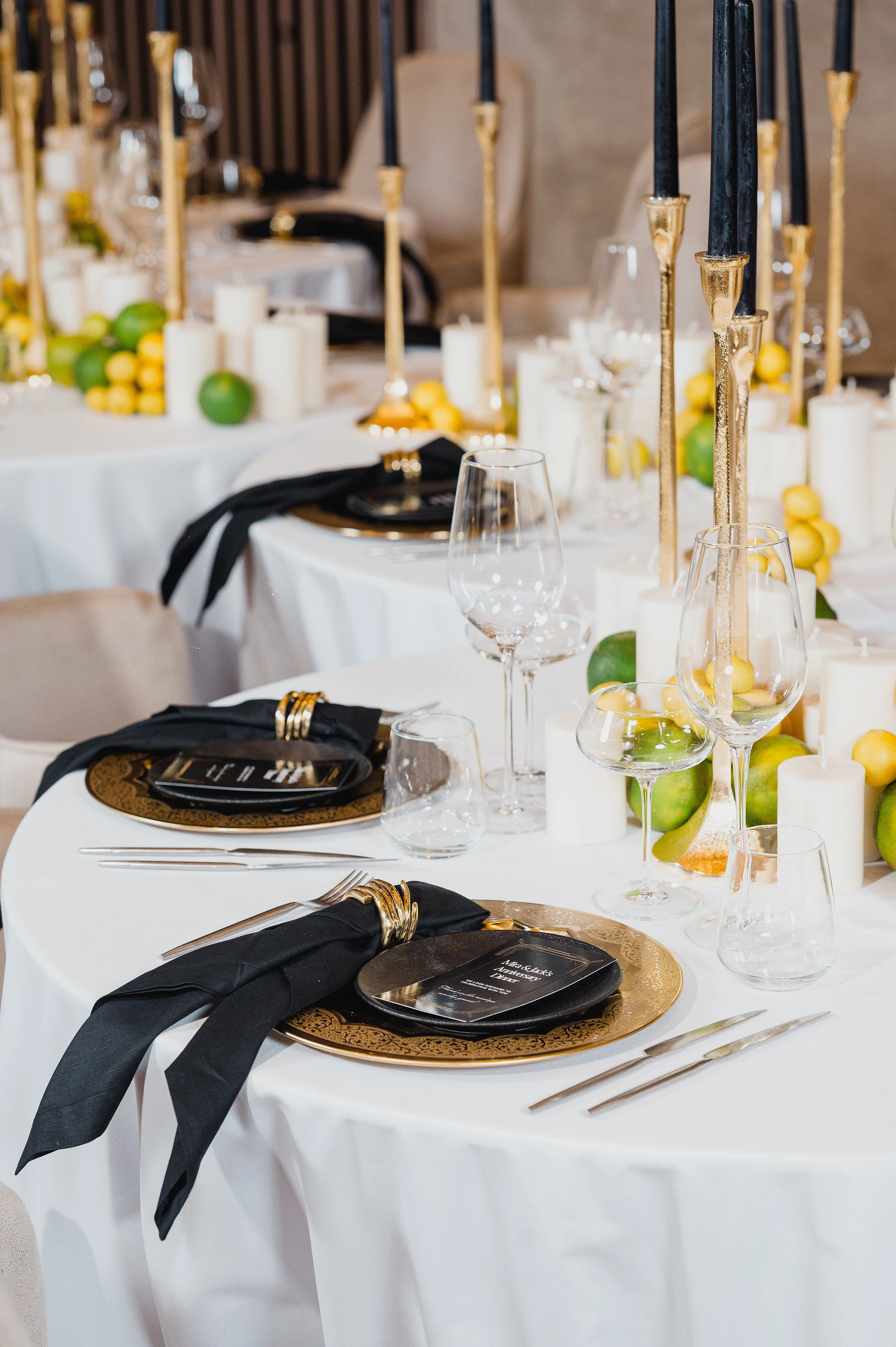 Elegant banquet table setting with black napkins, gold chargers, wine glasses, decorative candles, lemons, and limes for a formal event.