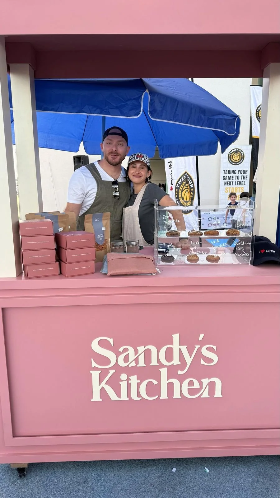 A man and woman smiling at a pink snack stand named 'Sandy's Kitchen'. They are behind a counter with boxes, jars, and a transparent display case with cookies. The stand is shaded by a blue umbrella, and there are banners and a sign in the background.