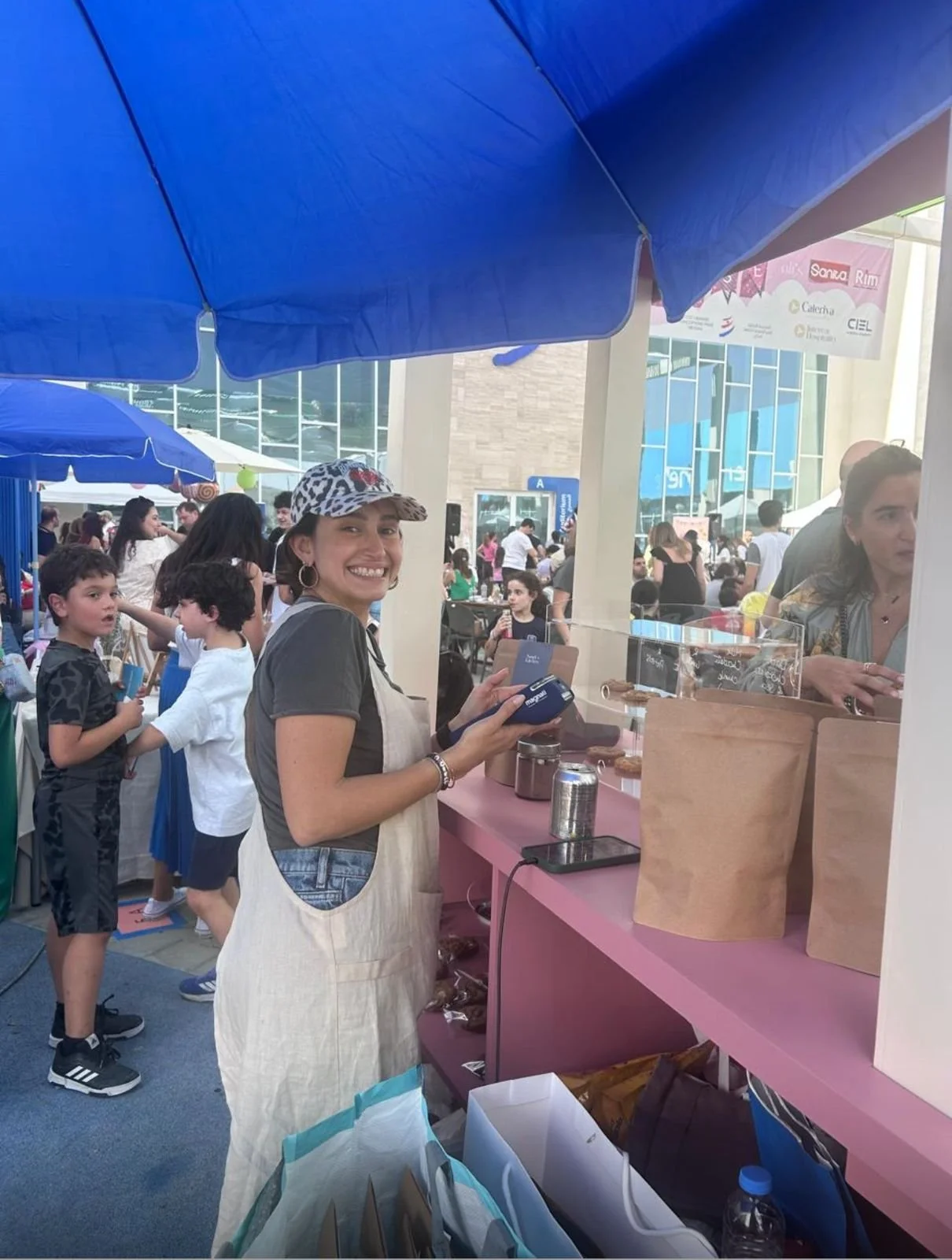 Smiling woman at a market stall, surrounded by people and blue umbrellas.