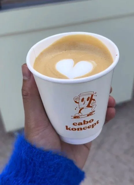 A hand holding a white cup of coffee with a heart-shaped foam art on top. The cup has a cartoon character and the text 'cabo koncept' printed on it.