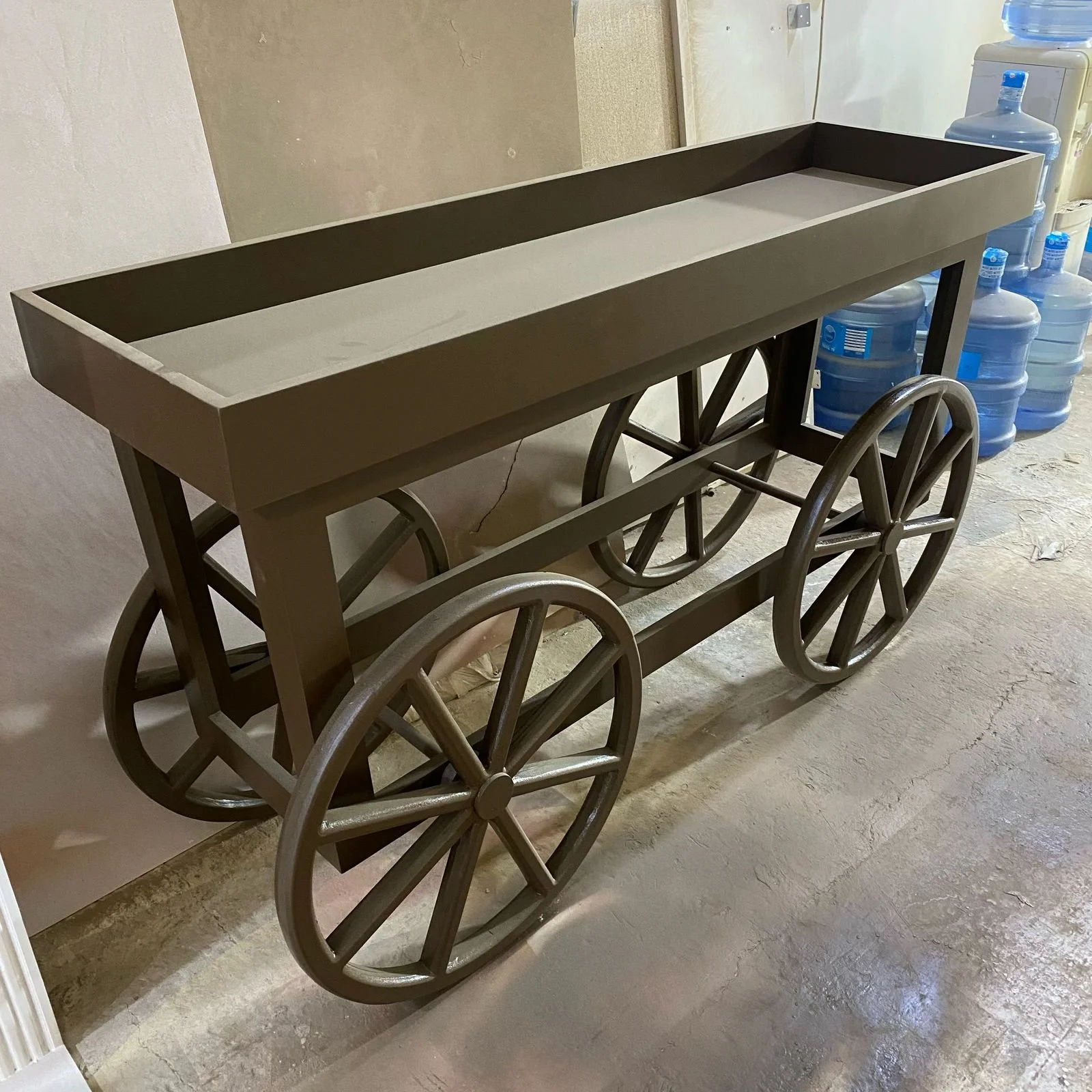 A vintage wooden cart with large wheels, painted in a neutral color, situated in a room with water dispensers in the background.