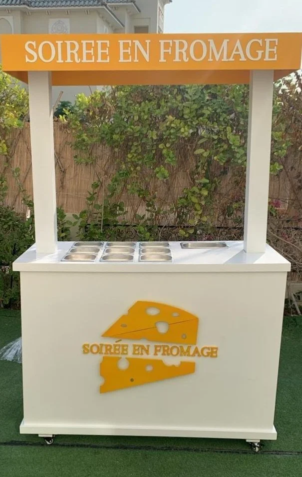 A white cheese station cart with a yellow cheese graphic and the words "SOIRÉE EN FROMAGE" on the front. The top has metal containers for serving cheese, and a yellow sign with the same phrase on it is above the station.