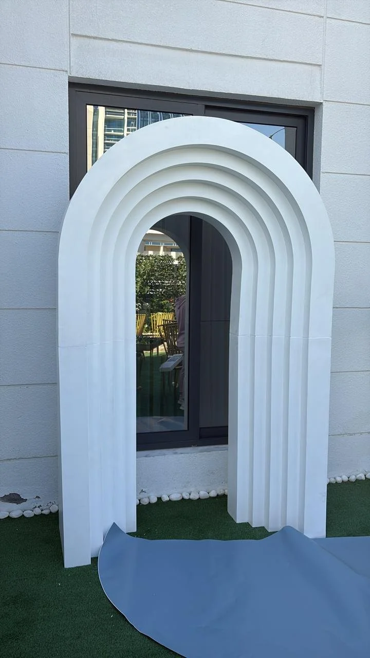 A white, arched, layered decorative structure standing outdoors in front of a window, with a blue mat or fabric at its base and a green artificial lawn surface.