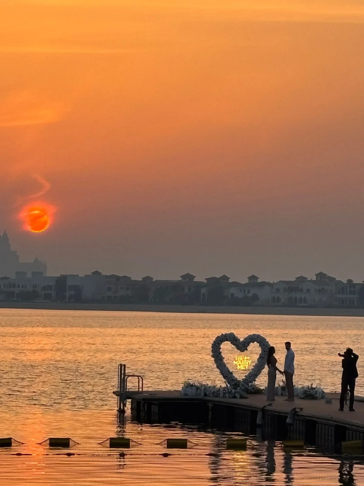 A couple is standing on a dock by the water at sunset, holding hands with a heart-shaped floral arch behind them that says 'Will You Marry Me' in lights. A photographer is capturing the moment on the right. The sky is orange with the sun low on the horizon.
