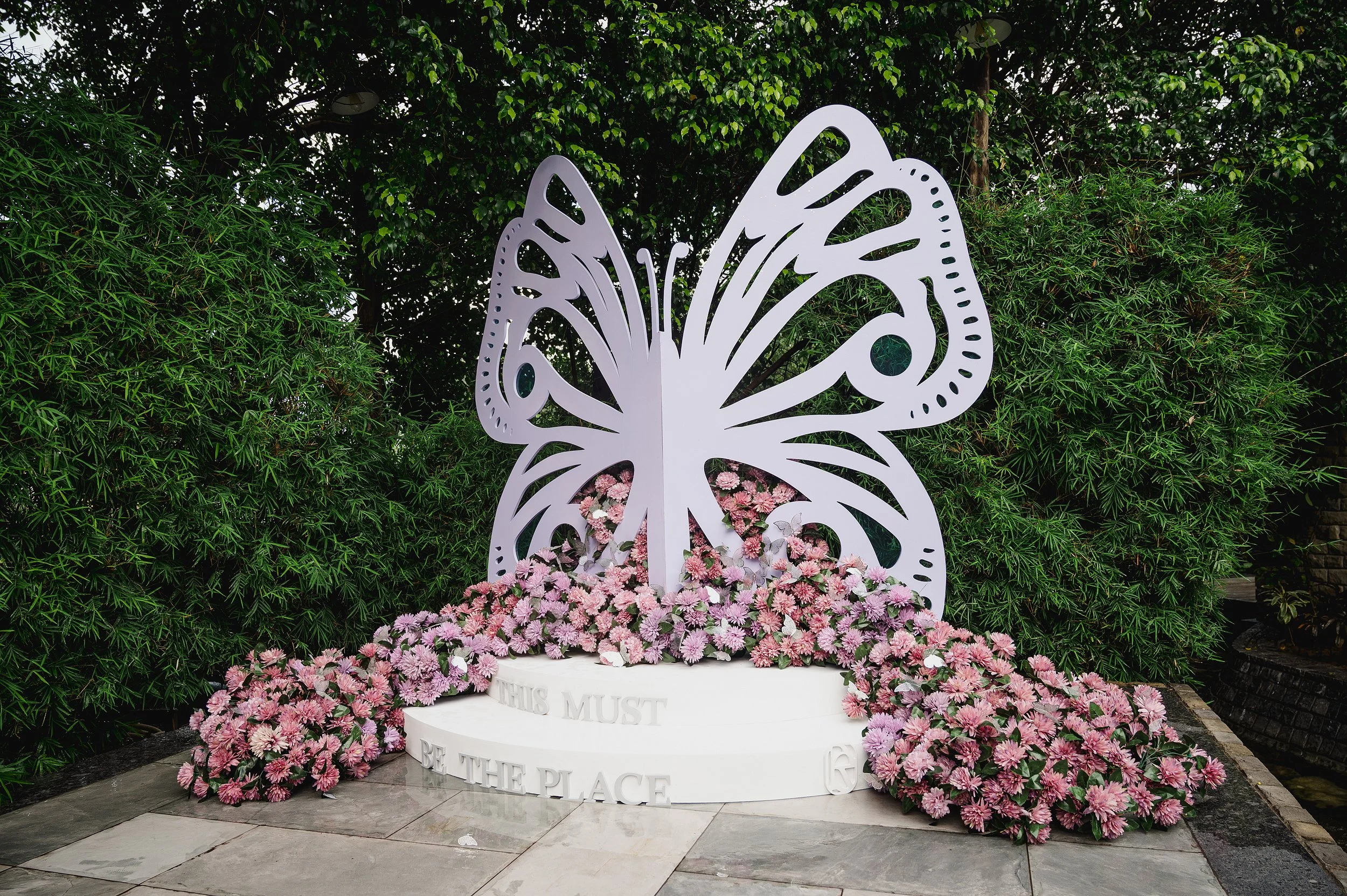 A large white butterfly sculpture surrounded by pink and purple flowers with green trees in the background.