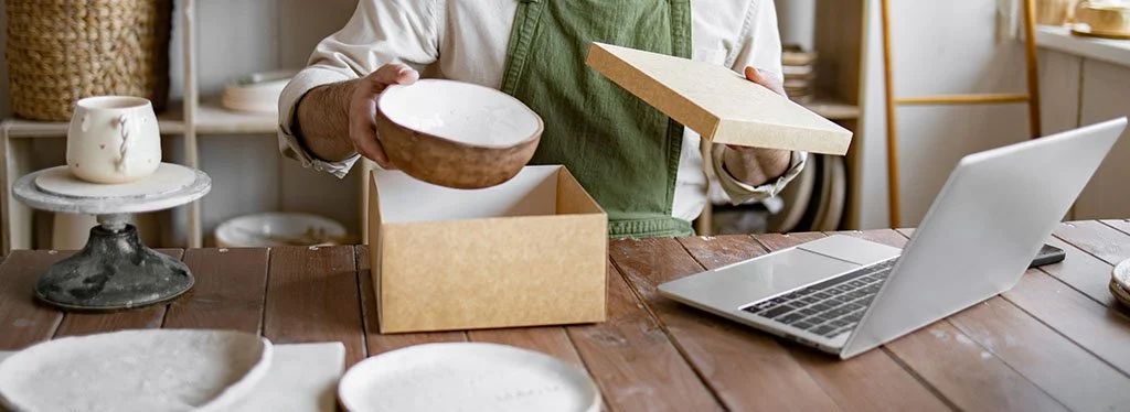 Person packing a ceramic bowl into a shipping box, with computer open on the table