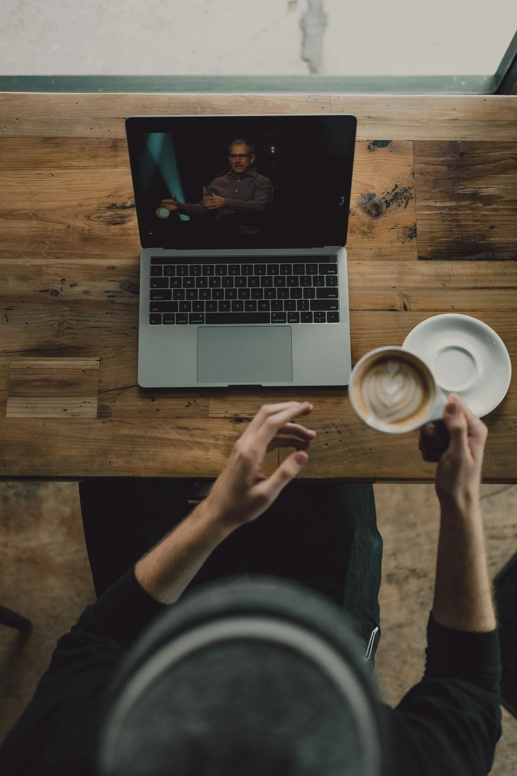 Person watching another person speaking on a laptop while holding a cup of coffee, sitting at a wooden table.