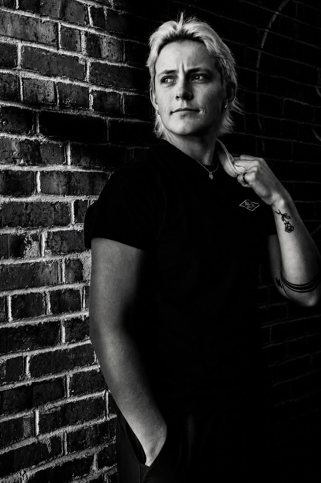 A woman with blonde hair standing against a brick wall, looking to the side with a serious expression, wearing a black shirt and tattoos on her arm.