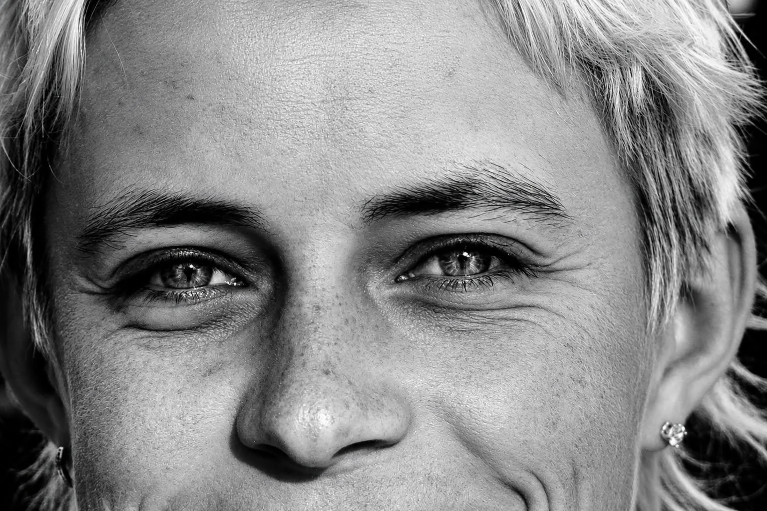 Close-up black and white photo of a smiling woman with short, light-colored hair and earrings.