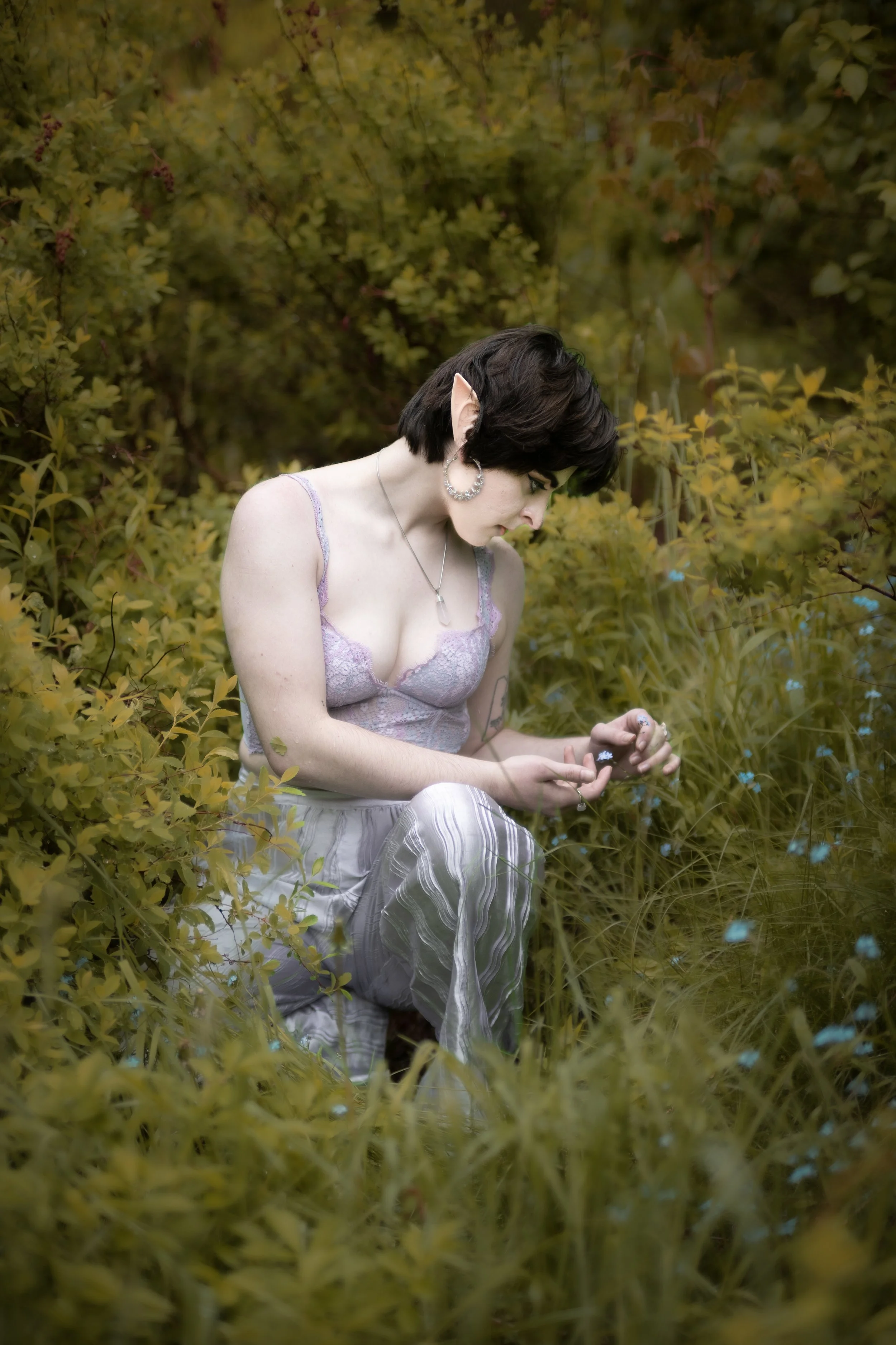 A person with elf ears and short dark hair kneels among green and yellow foliage, looking at or holding small objects in their hands.