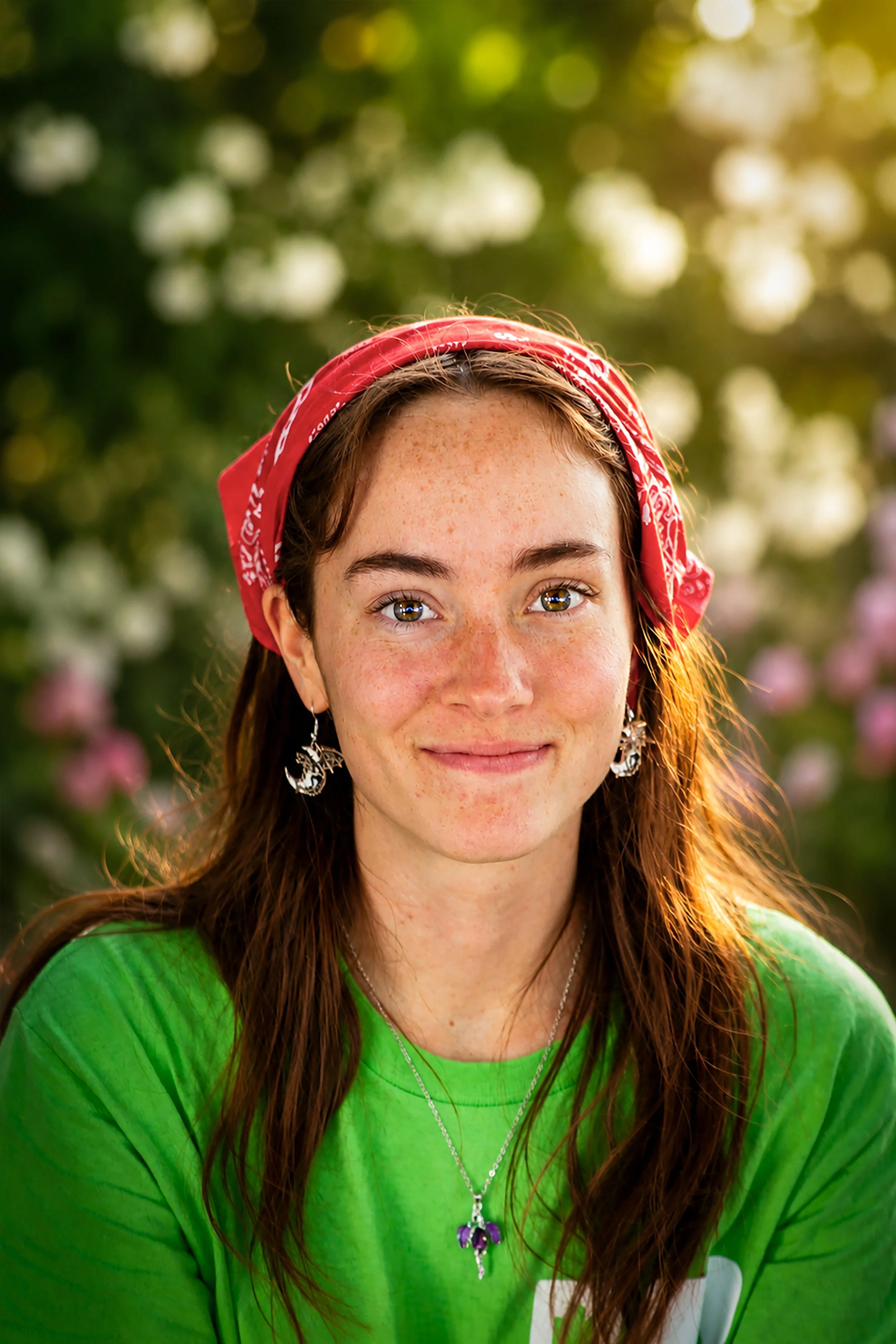 Close-up of a young woman with long, auburn hair wearing a red bandana on her head, a green shirt, and silver accessories, outdoors with blurred greenery and flowers in the background.